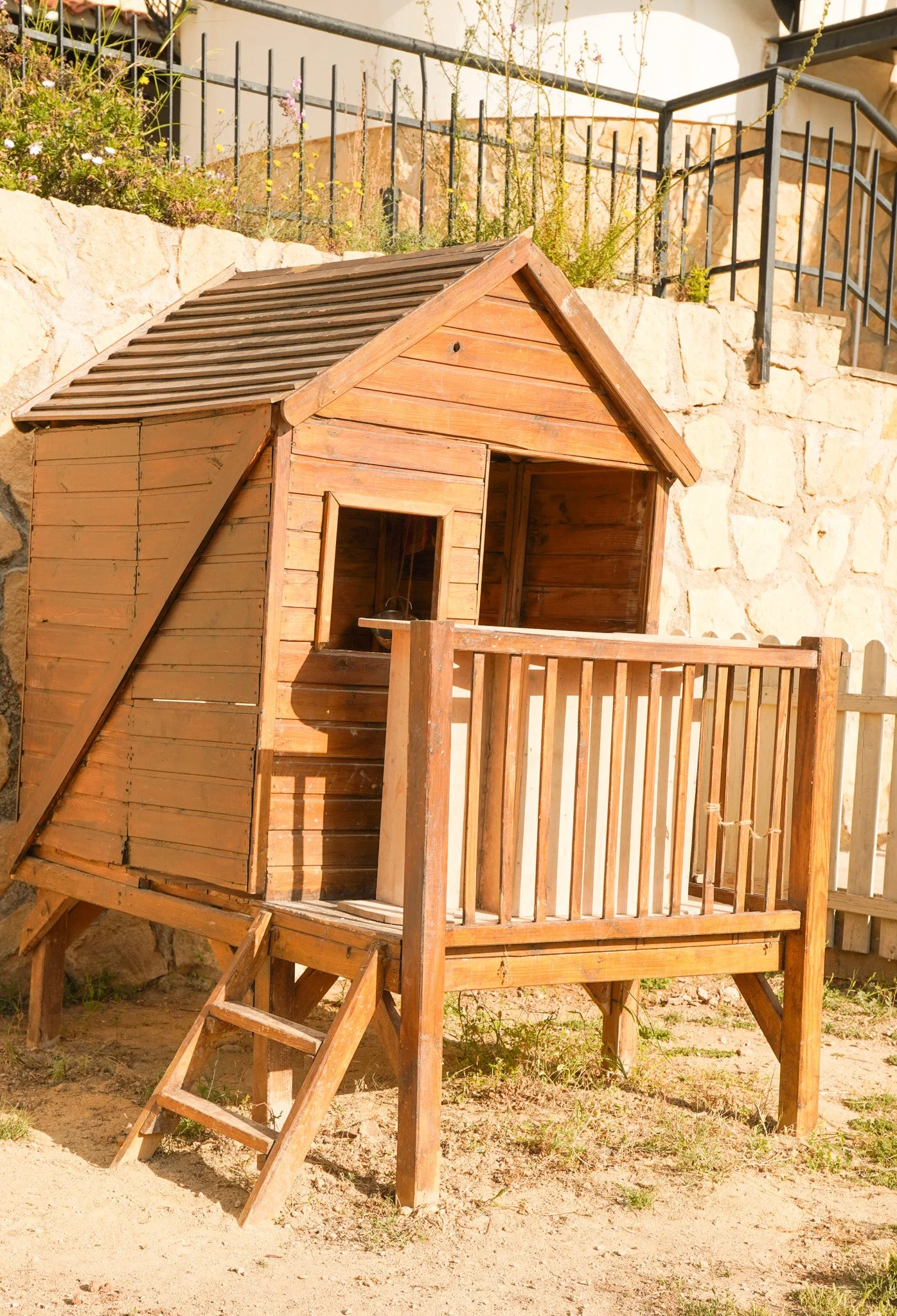 Casa de madera en tonos naturales con escalera en el frente y barandales, situada en un entorno con muro de piedra y vegetación.