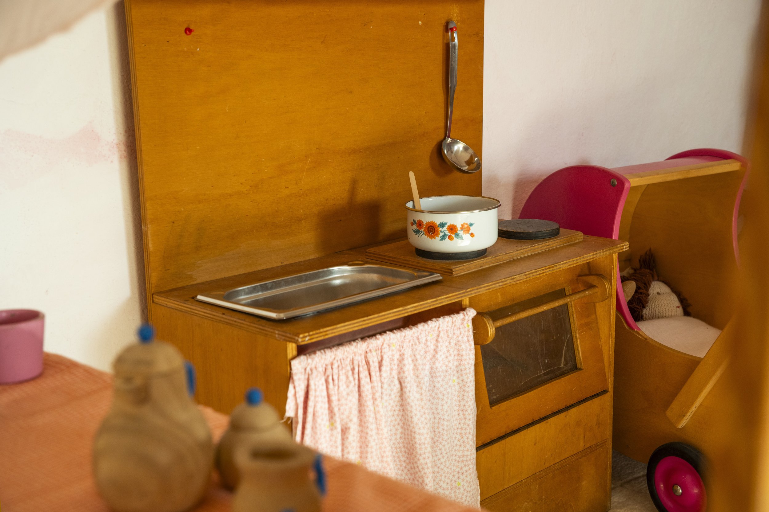 Mueble de cocina de madera con un recipiente con flor en la parte superior, un cucharón y una bandeja de metal. Incluye toallas de mano colgadas y detalles de color rosa en el entorno.