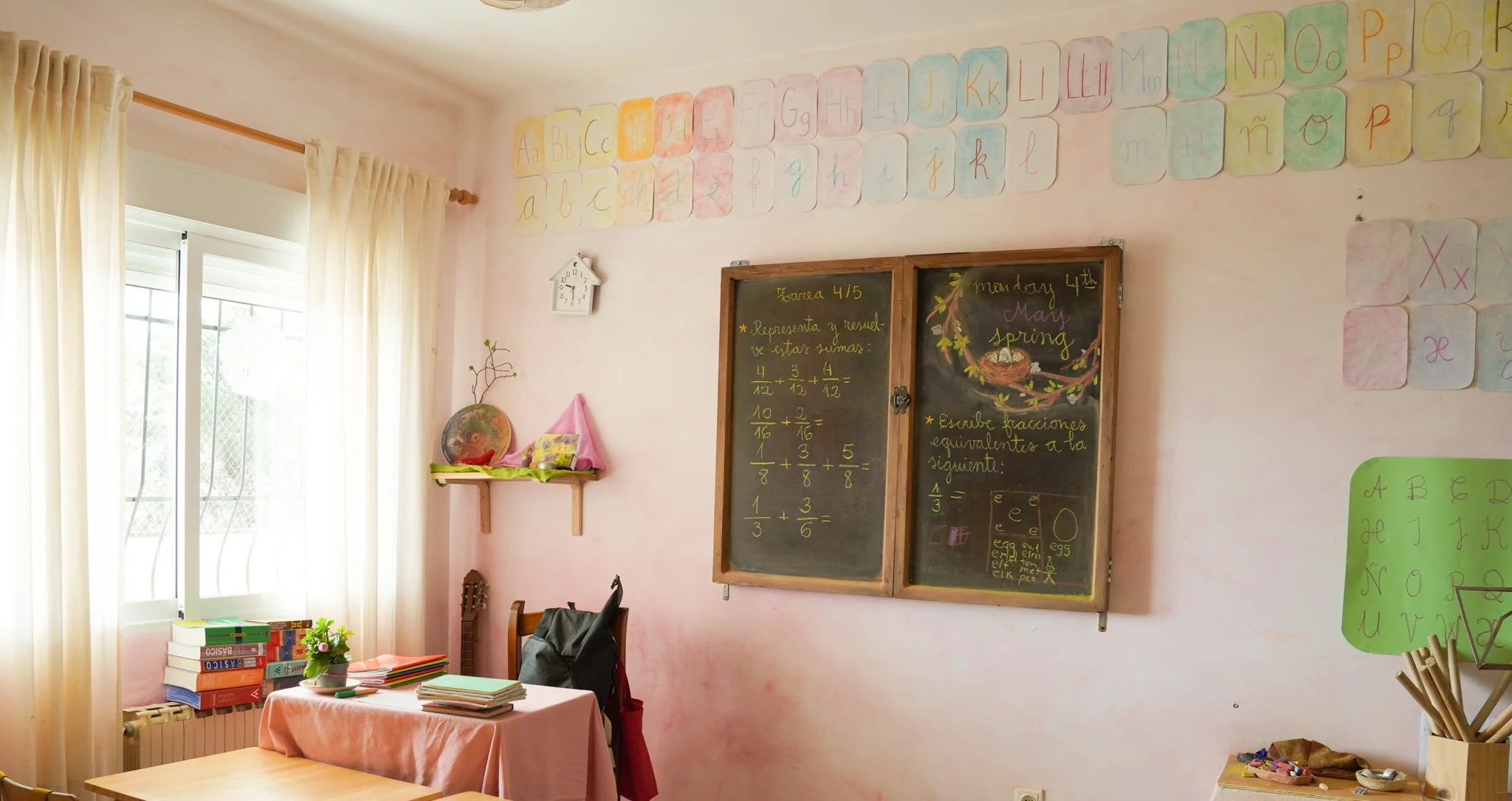Sala de clases con paredes rosadas, decoradas con letras del abecedario en colores pastel. Hay una pizarra con ejercicios de suma escritos en color amarillo y una lista de palabras en color rosa y amarillo. Ventana con cortinas blancas. Mesa con pila