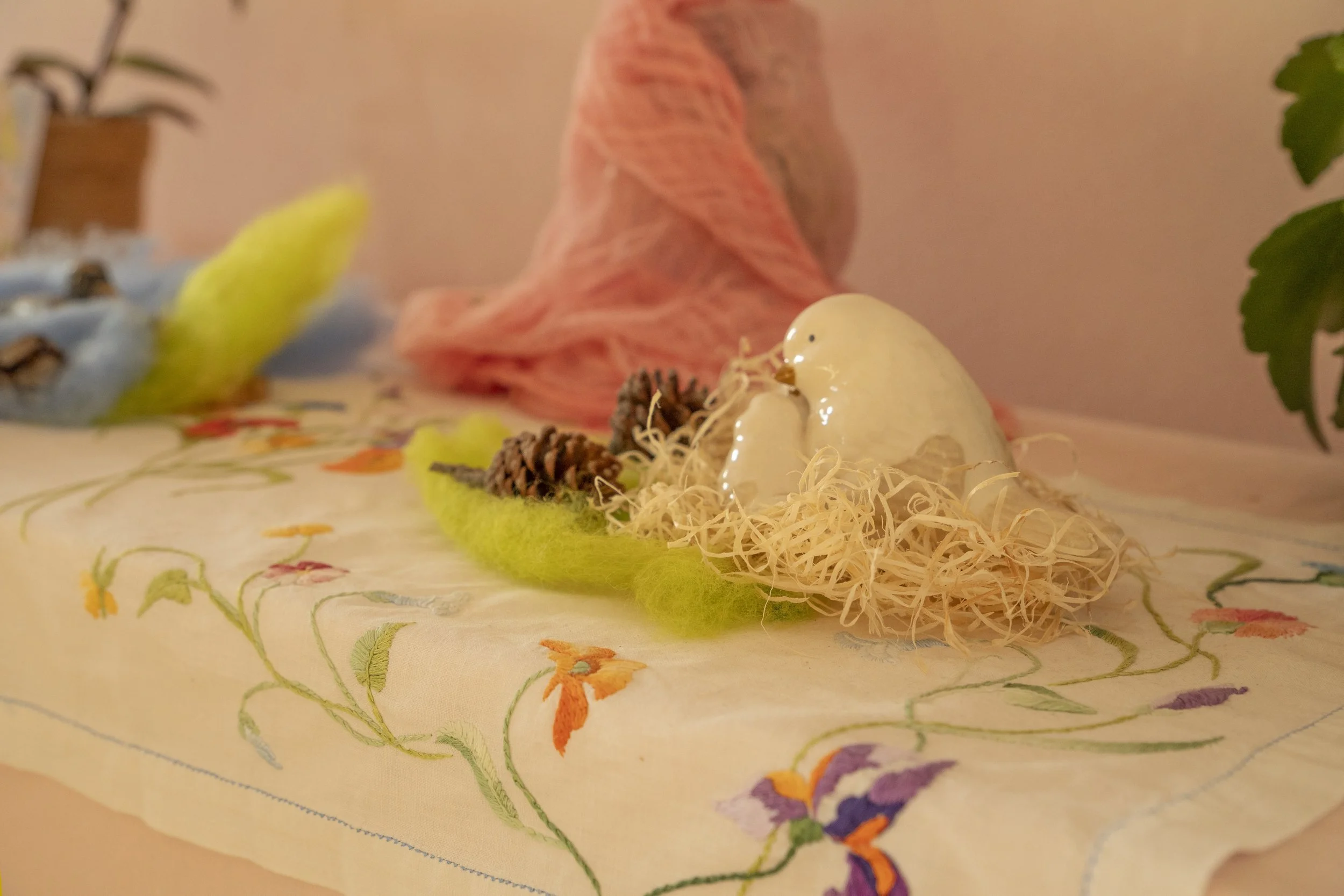 Una figura de cerámica en forma de pato en un entorno decorativo con plantas y una toalla con diseño floral sobre una mesa.