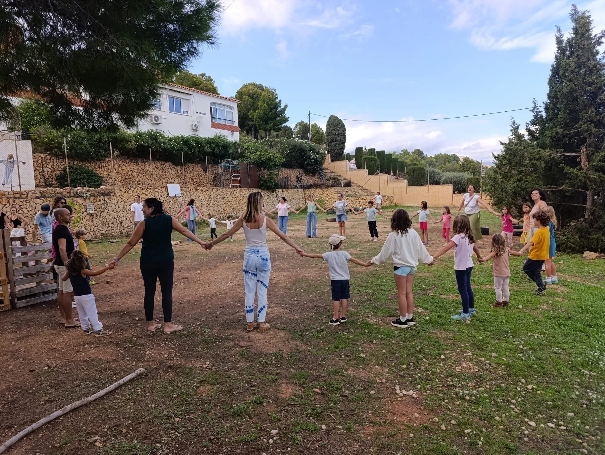 Grupo de niños y adultos formando una cadena, jugando y sosteniéndose de las manos en un espacio al aire libre con árboles y una vivienda en el fondo.