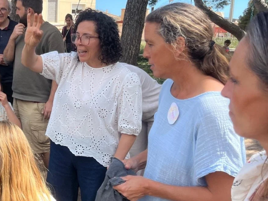 Mujer con gafas y blusa blanca hablando y gesticulando con la mano, rodeada de varias personas en un parque al aire libre.