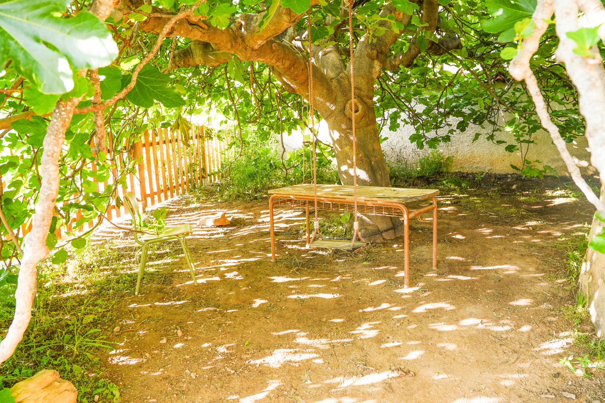 A columpio de madera suspendido de un árbol grande y frondoso en un jardín con malla y dos sillas de plástico verde y una cerca de madera en el fondo.