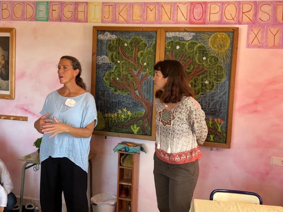 Dos mujeres conversan frente a un pizarrón con arbol y el sol, en un salón de clases decorado con letras del abecedario en la parte superior en colores pastel.