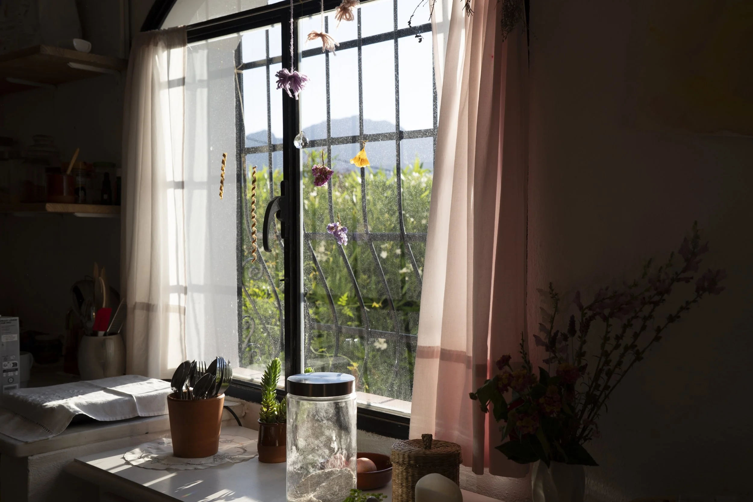 Vista de una ventana con cortinas beige y adornos colgados, con plantas en una mesa y una vista de montañas y vegetación en el exterior.