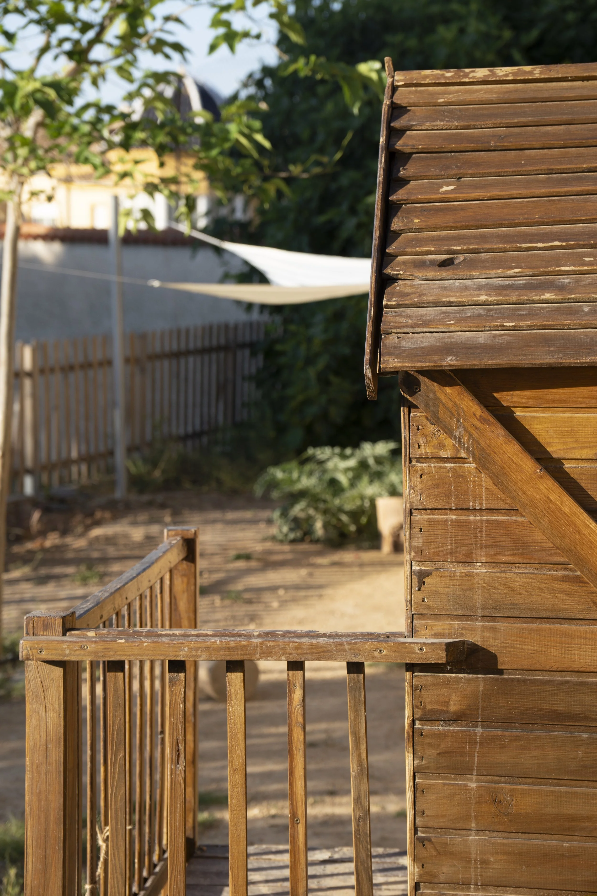 Vista de una cerca de madera y una colina o jardín con árboles y casa en el fondo, en un día soleado.