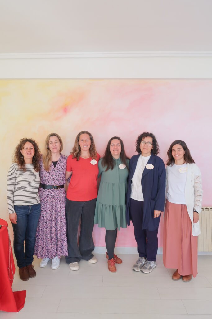 Seis mujeres de diferentes edades posando juntas en un fondo de pared coloreada con tonos pastel, en un ambiente interior.
