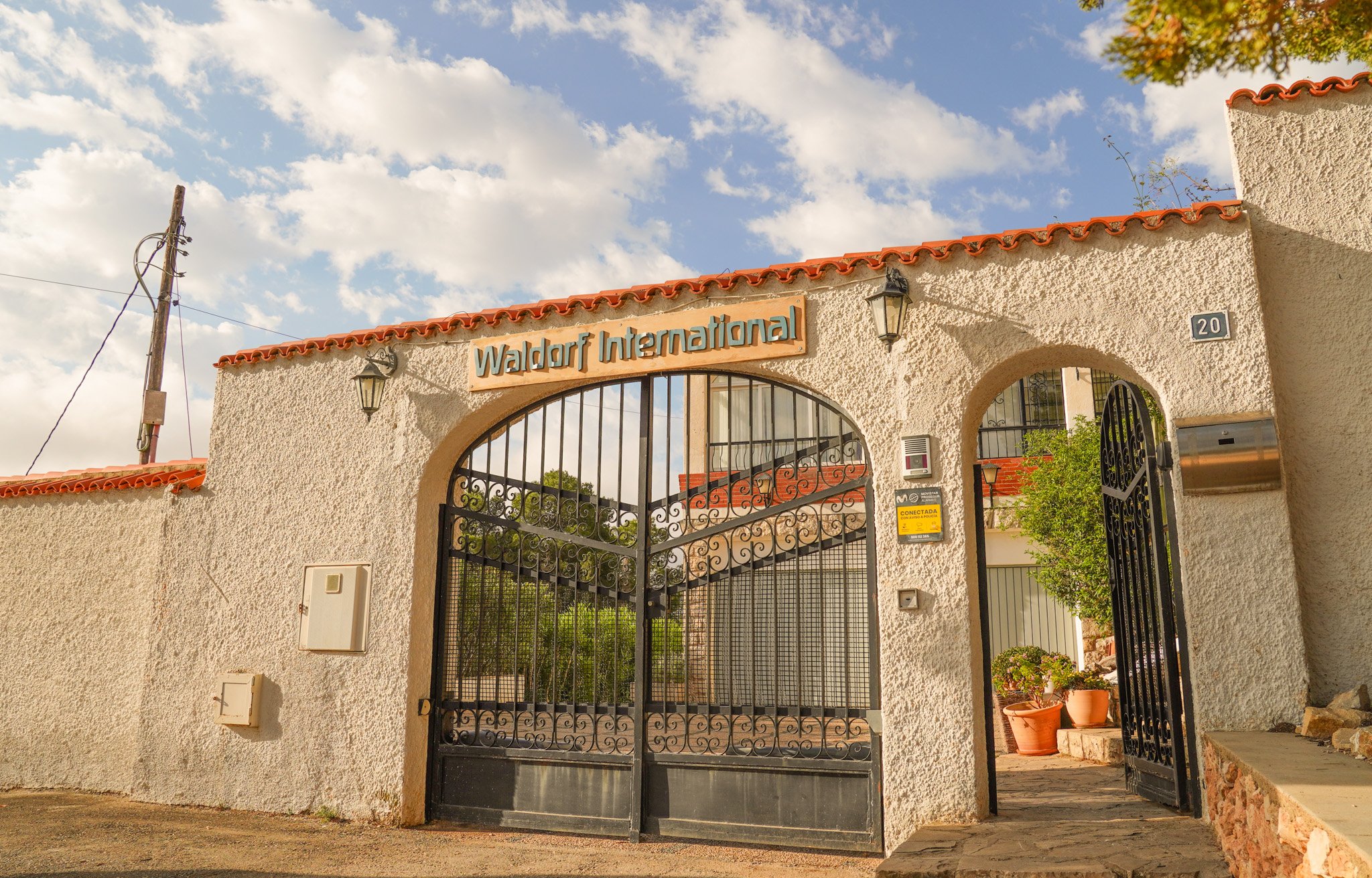 Entrada de una propiedad con reja de hierro negra y muro de textura blanca, señal que dice 'Waldorf International', pórtico con tejas rojas y una casa al fondo. Hay un poste de electricidad y plantas en macetas.