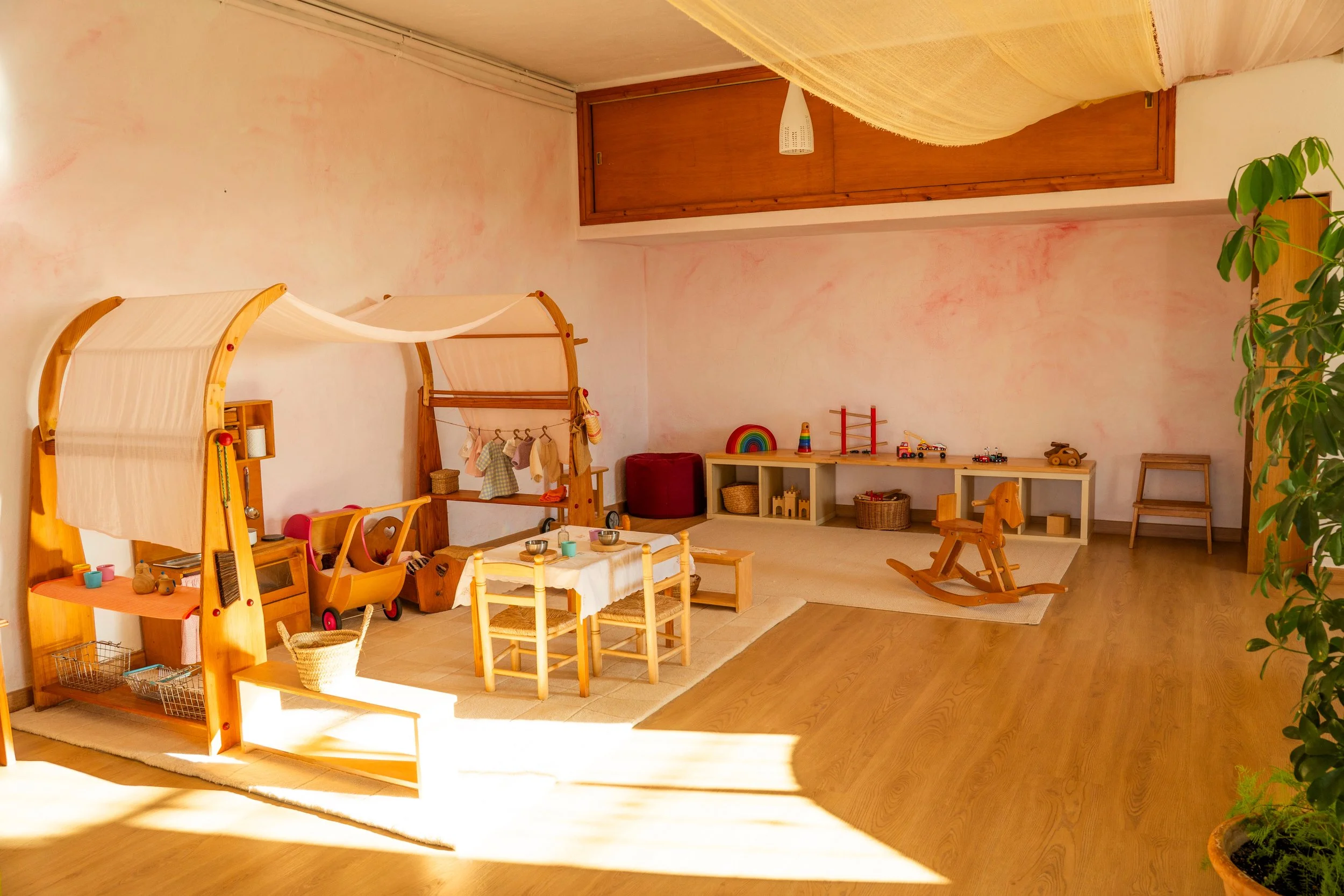 Habitación infantil con juguetes de madera, mesa de juego, y muebles de madera, paredes rosadas, y una planta verde en la esquina.