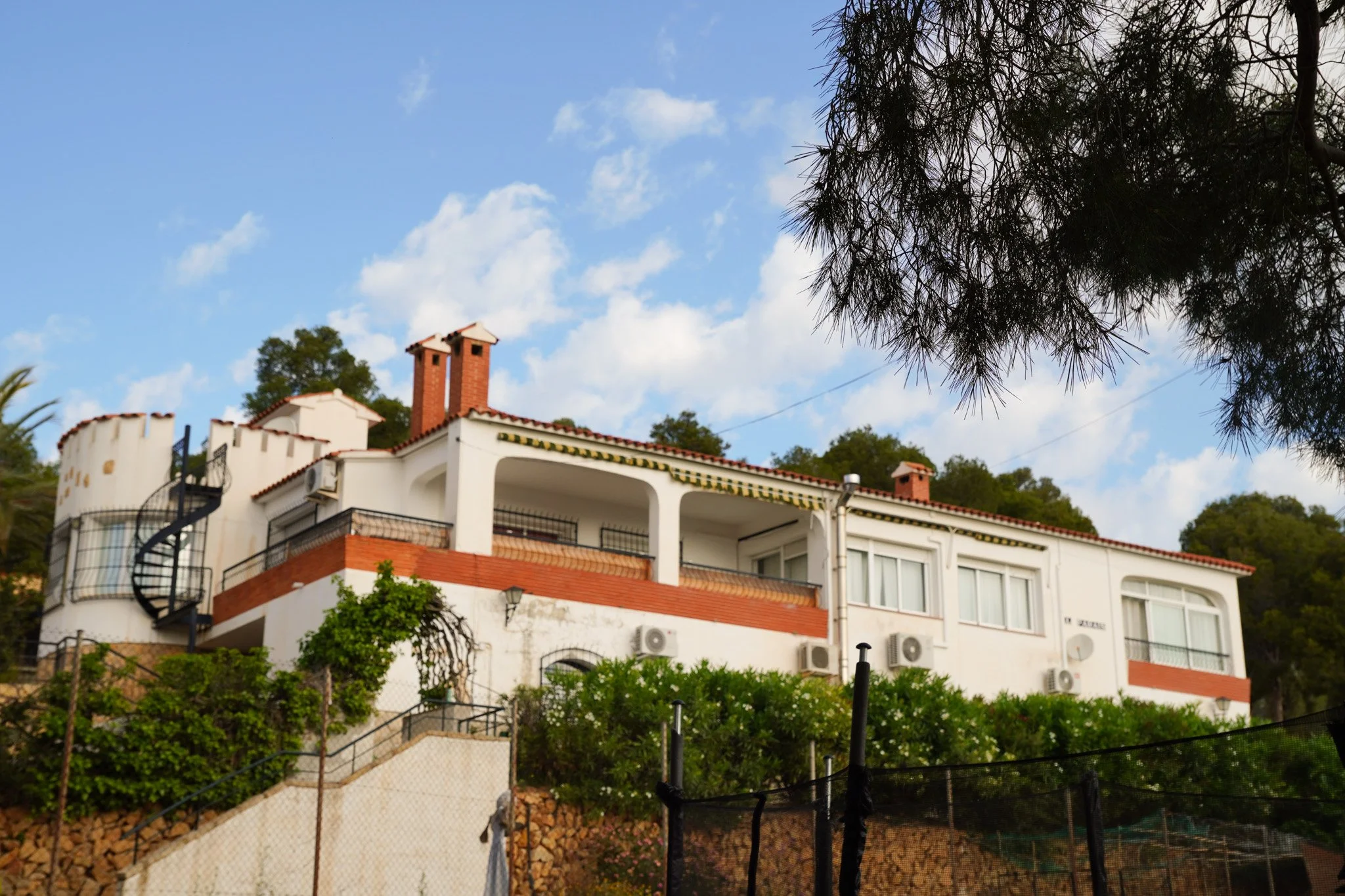 Casa blanca de múltiples niveles con azoteas y chimeneas rojas, rodeada de árboles y un cielo azul con nubes en el fondo.
