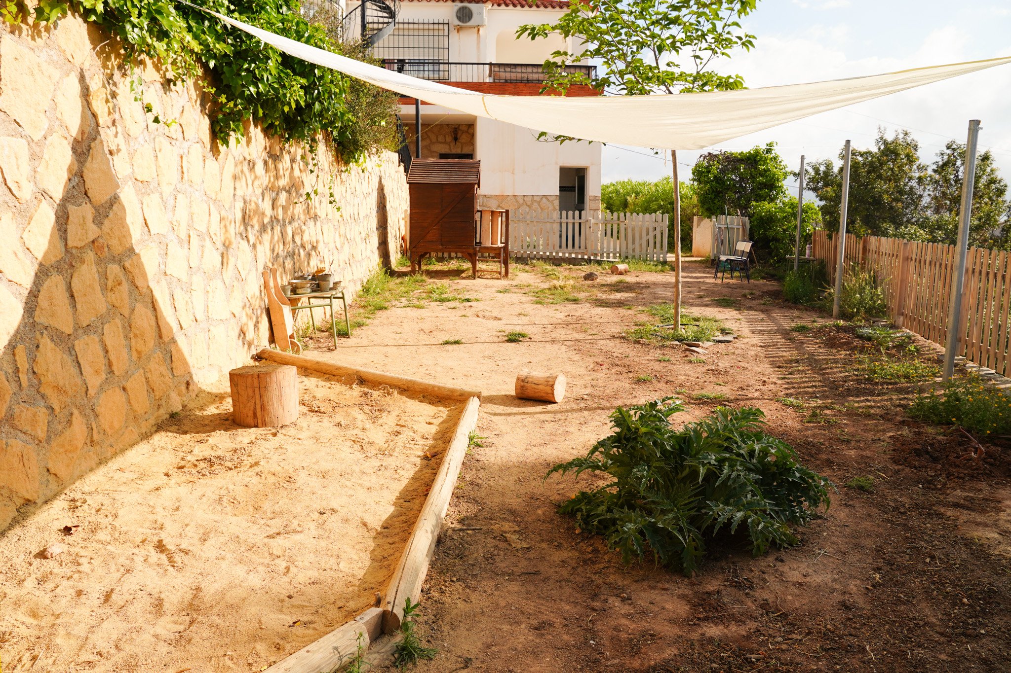 Un patio con suelo de tierra, plantas verdes, una barda de piedra y una cerca de madera, con sombra de una tela blanca, en un día soleado.
