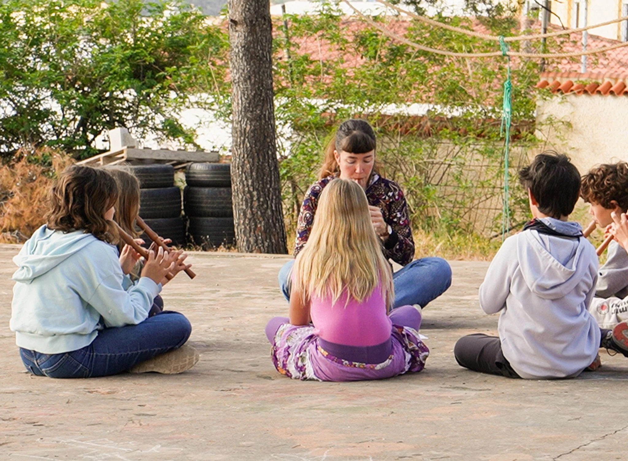Grupo de niños y un adulto participando en una actividad al aire libre, sentados en círculo sobre el suelo en un espacio rodeado de árboles y plantas.