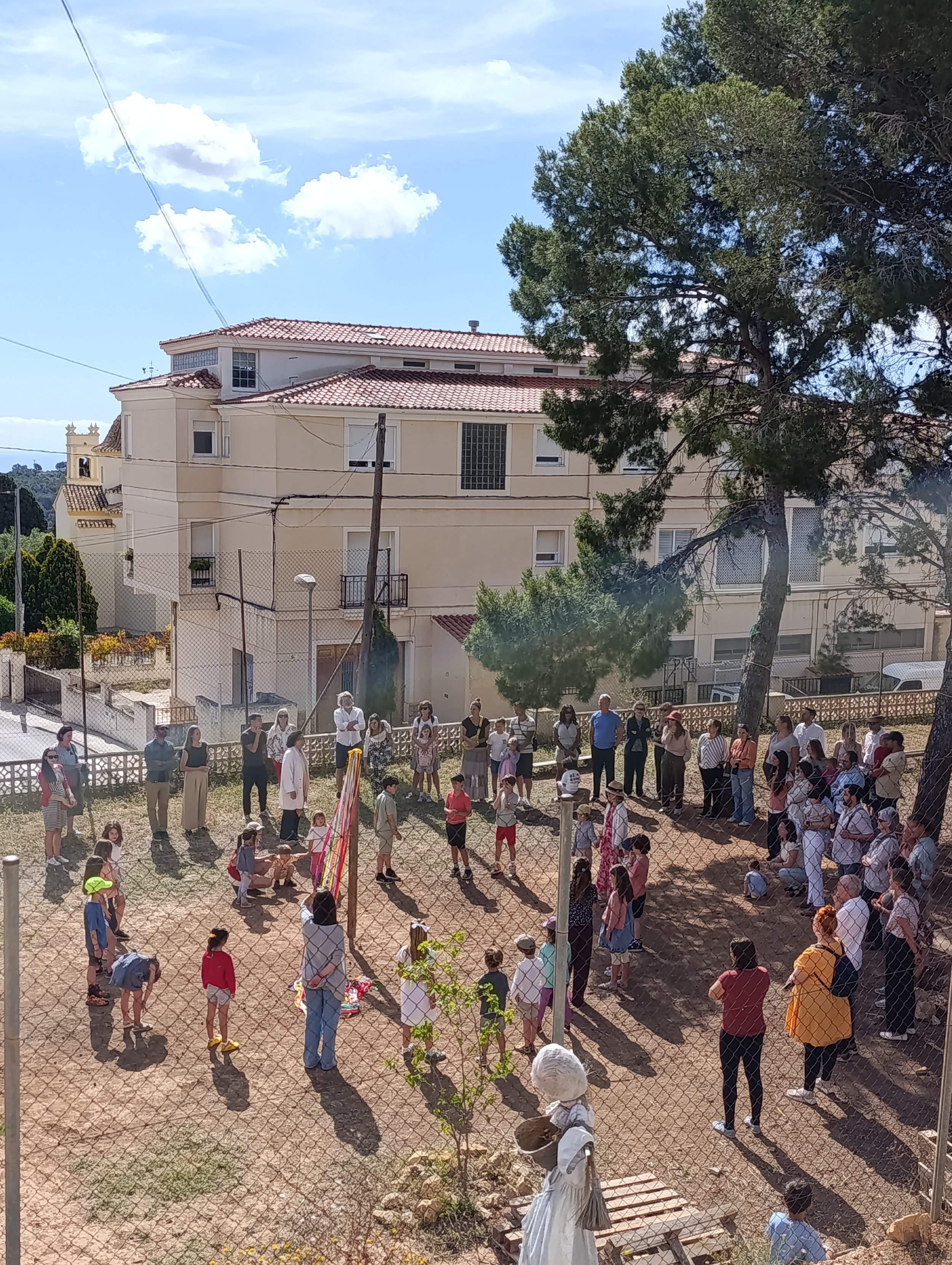 Una multitud de personas se encuentra en un espacio abierto cercado, algunos están conversando y otros miran en diferentes direcciones, rodeados de árboles y una casa de varios pisos con techos de tejas rojas al fondo.