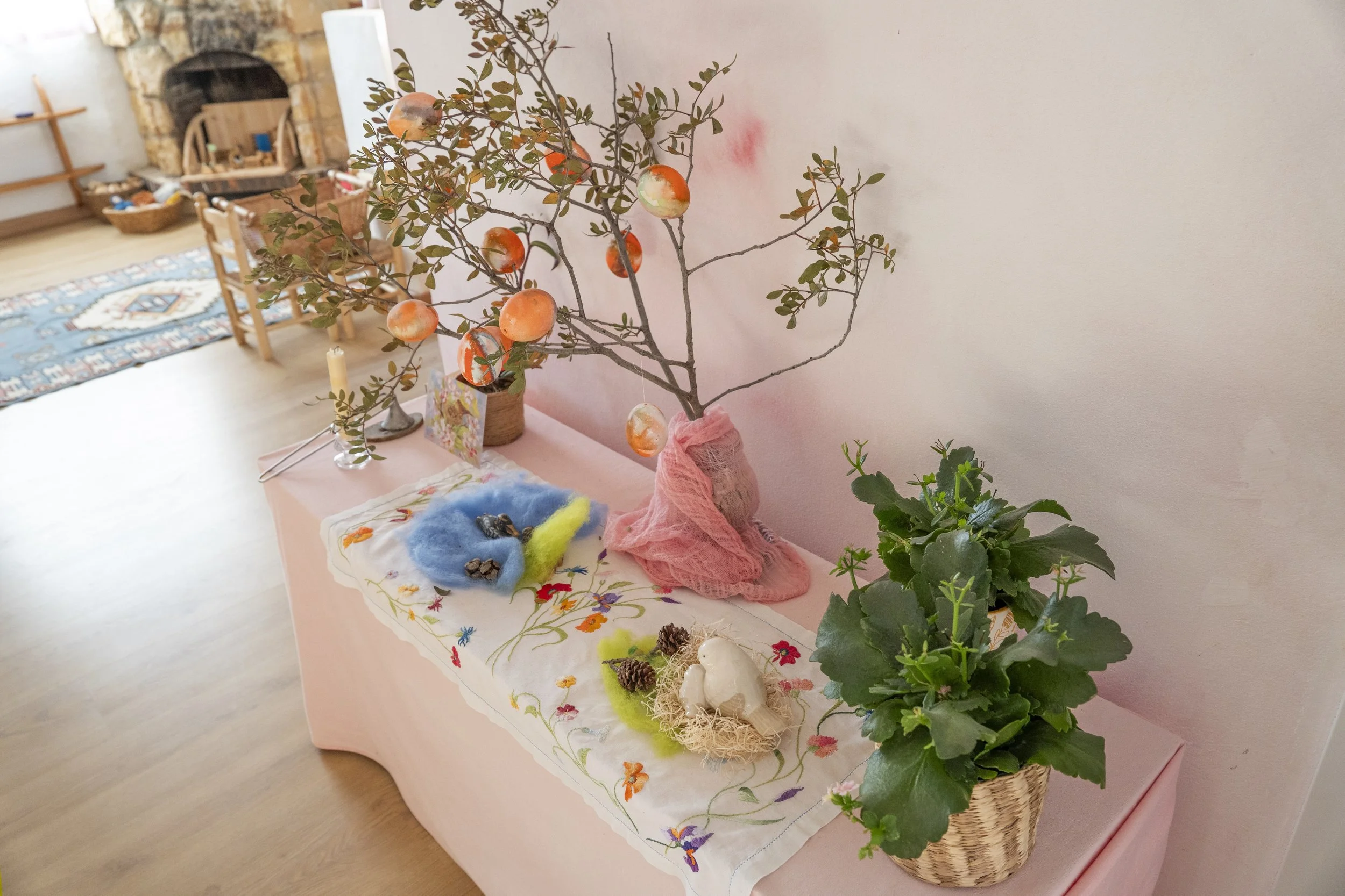 Una mesa con una decoración de temática de otoño o vacaciones, con un árbol artificial con calabazas decorativas colgantes, plantas verdes en macetas, objetos de fantasía como ovejas de peluche, piñas y huevos decorativos. La mesa está cubierta con u