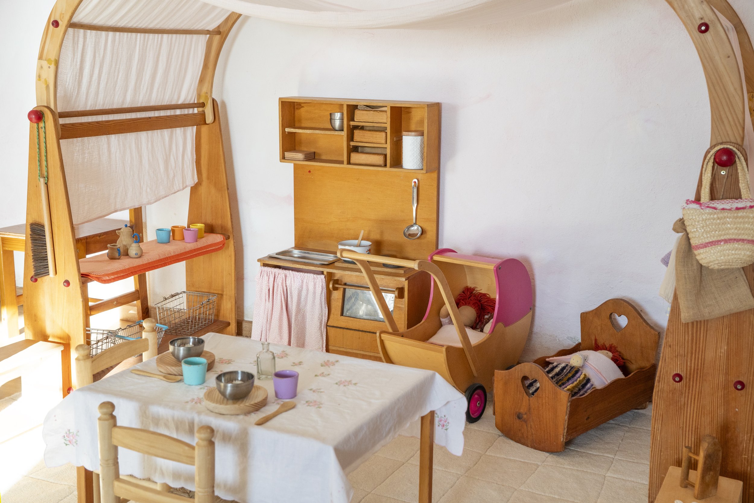 Habitación infantil con muebles de madera, cama en forma de carro, estanterías, juguetes, y mesa con platos y vasos de colores.