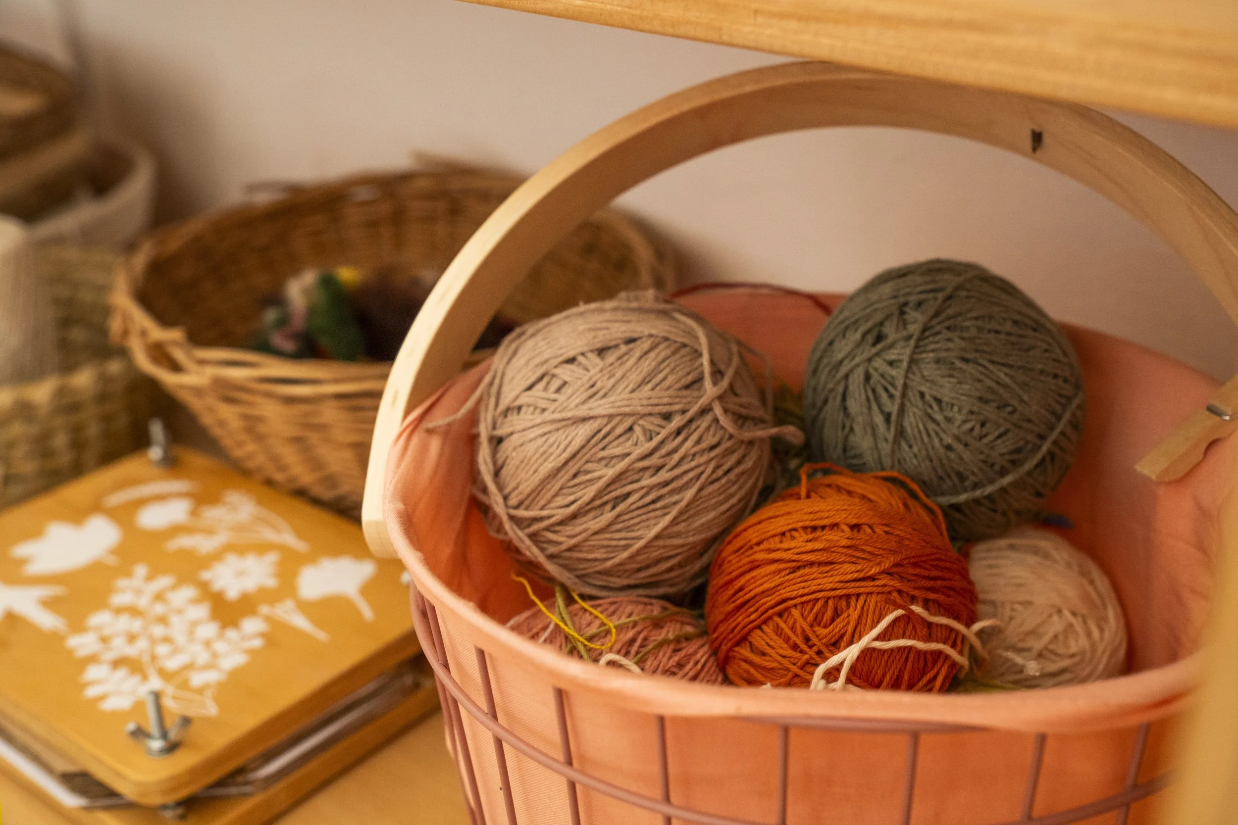 Un cesto de hilo de colores en tonos beige, gris y naranja, con una estructura de metal color cobre, en un entorno decorativo con cestas de mimbre y manteles con patrones.
