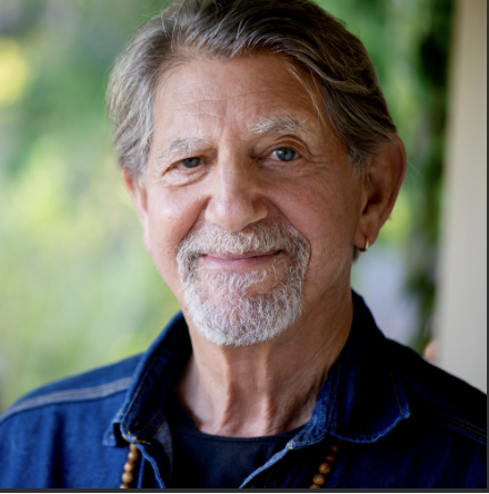 Close-up of an elderly man with gray hair, beard, and mustache, wearing a denim jacket and smiling, outdoors with a blurred green background.