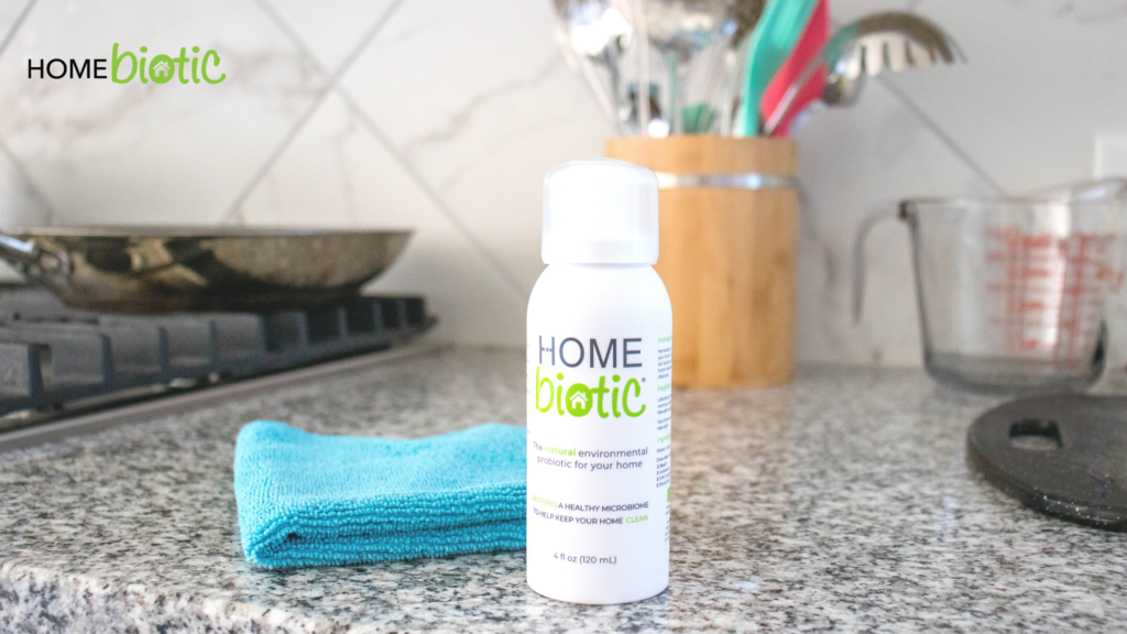 A bottle of Home Biotic probiotic in a kitchen with a blue cloth, a utensil holder, a measuring cup, and a stove in the background.