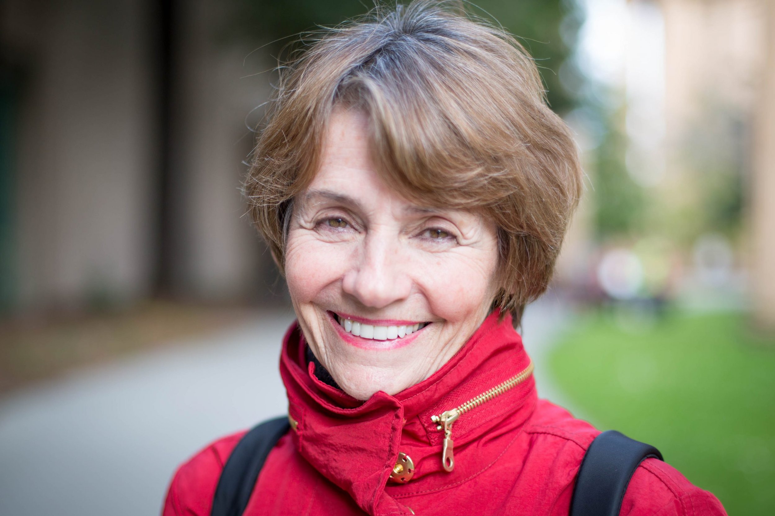 Smiling woman with short brown hair wearing a red jacket with black backpack strap outdoors.