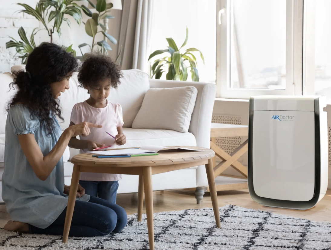 A woman and a young girl sitting on the floor at a small wooden table, looking at a notebook with colored pens, with a window and houseplants in the background, and an air purifier labeled 'AIR Doctor' nearby.