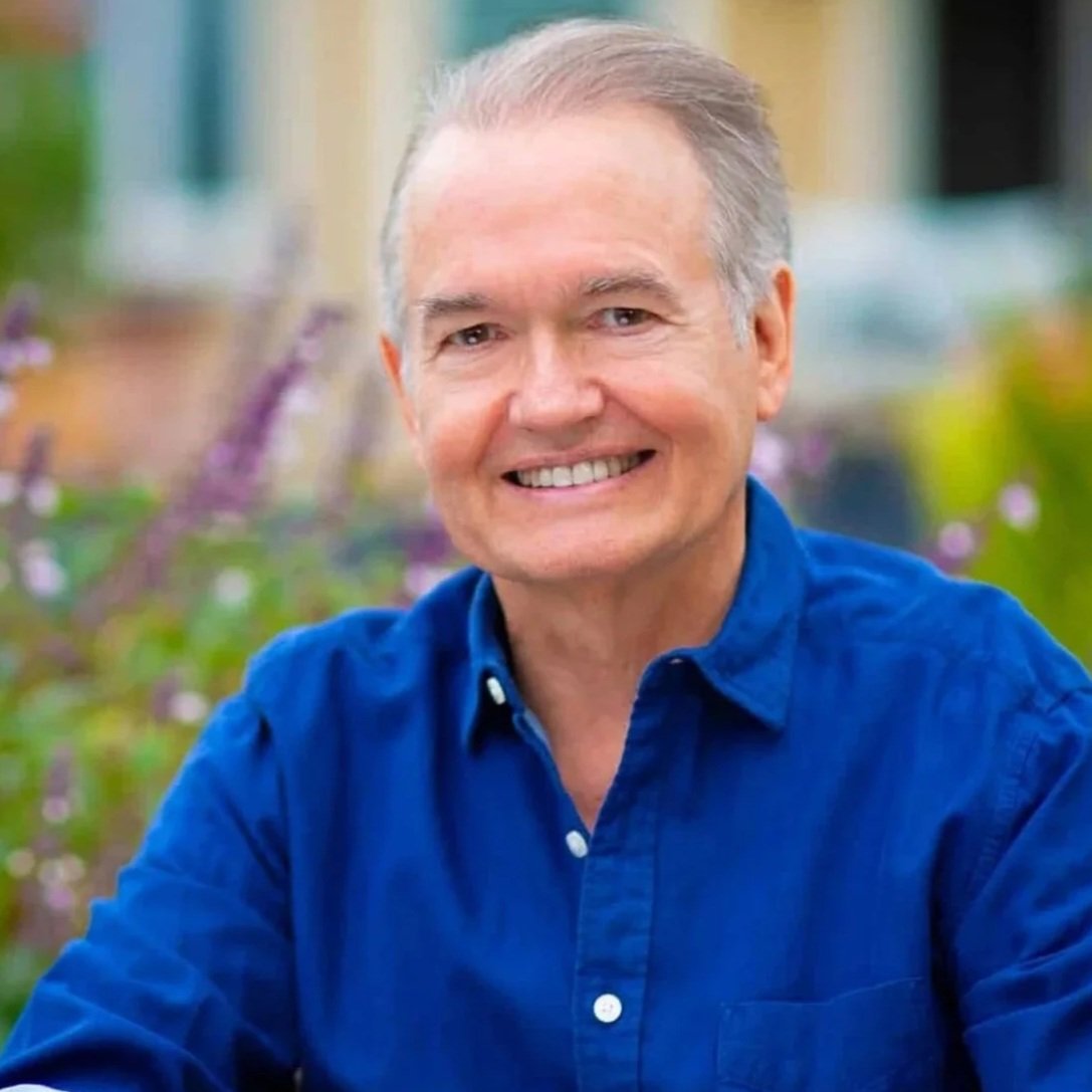 Smiling middle-aged man with short gray hair wearing a blue collared shirt, outdoors with a blurred background of greenery and flowers.