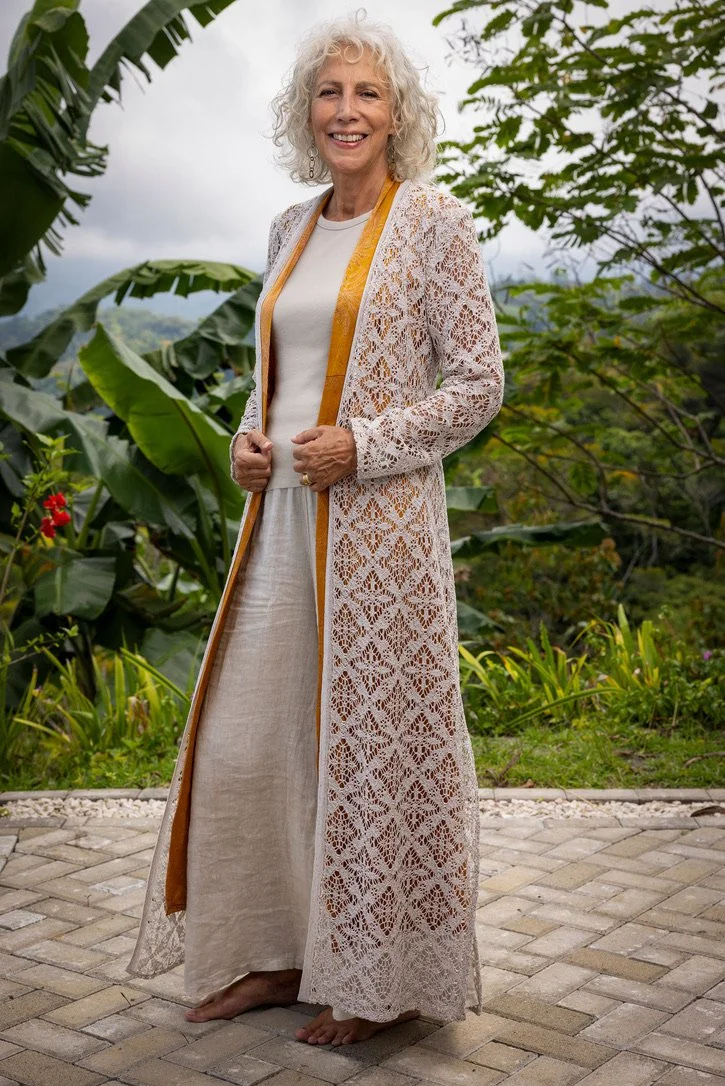 Older woman with curly gray hair standing outdoors on a brick path, surrounded by lush green tropical plants, smiling in a long white lace dress and a beige skirt, with a lightweight, open, embroidered cardigan.