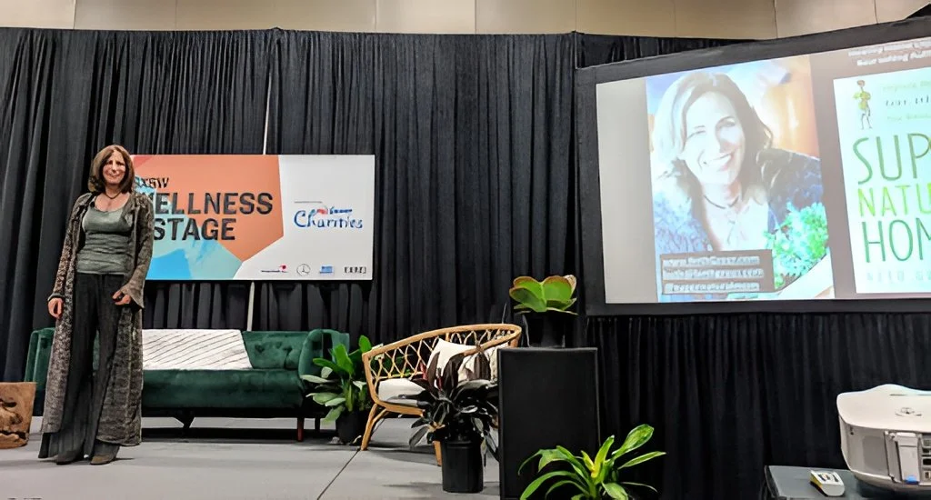 Woman standing on stage with black curtains, green sofa, and potted plants, at wellness event with presentation and banner.