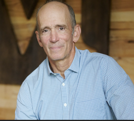 A middle-aged man wearing a blue checkered shirt, sitting indoors with wooden wall decor in the background.