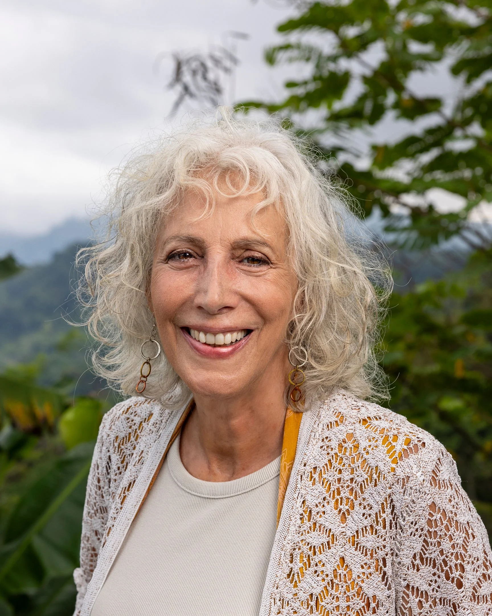 Close-up of smiling elderly woman with curly gray hair wearing earrings and a cream-colored crochet cardigan, standing outdoors with green leaves and mountains in the background.