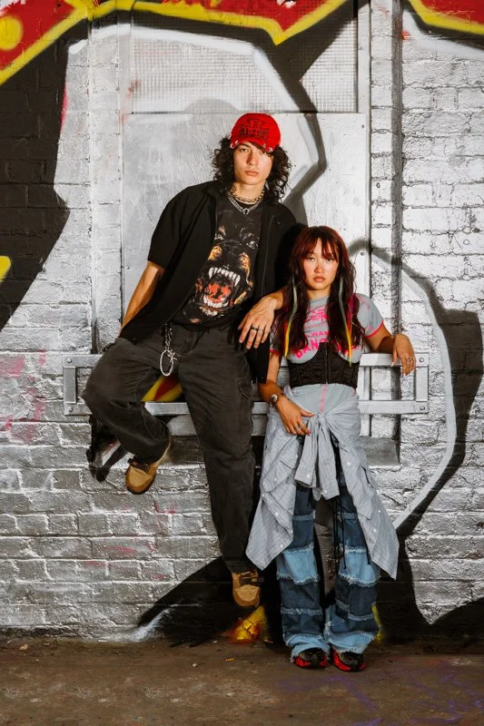 Two young people posing in front of a graffiti-covered brick wall. The person on the left is sitting on a ledge, wearing a red cap, black shirt with a graphic print, black pants, and tan shoes. The person on the right is standing, wearing a gray T-shirt with pink writing, a black vest, ripped blue jeans with a tied sweatshirt around the waist, and red shoes.