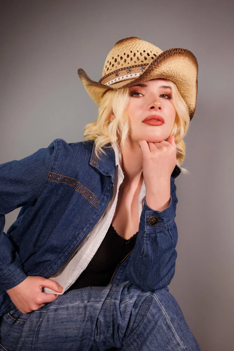Woman wearing a cowboy hat, denim jacket, and jeans, sitting and resting her chin on her hand, looking at the camera.