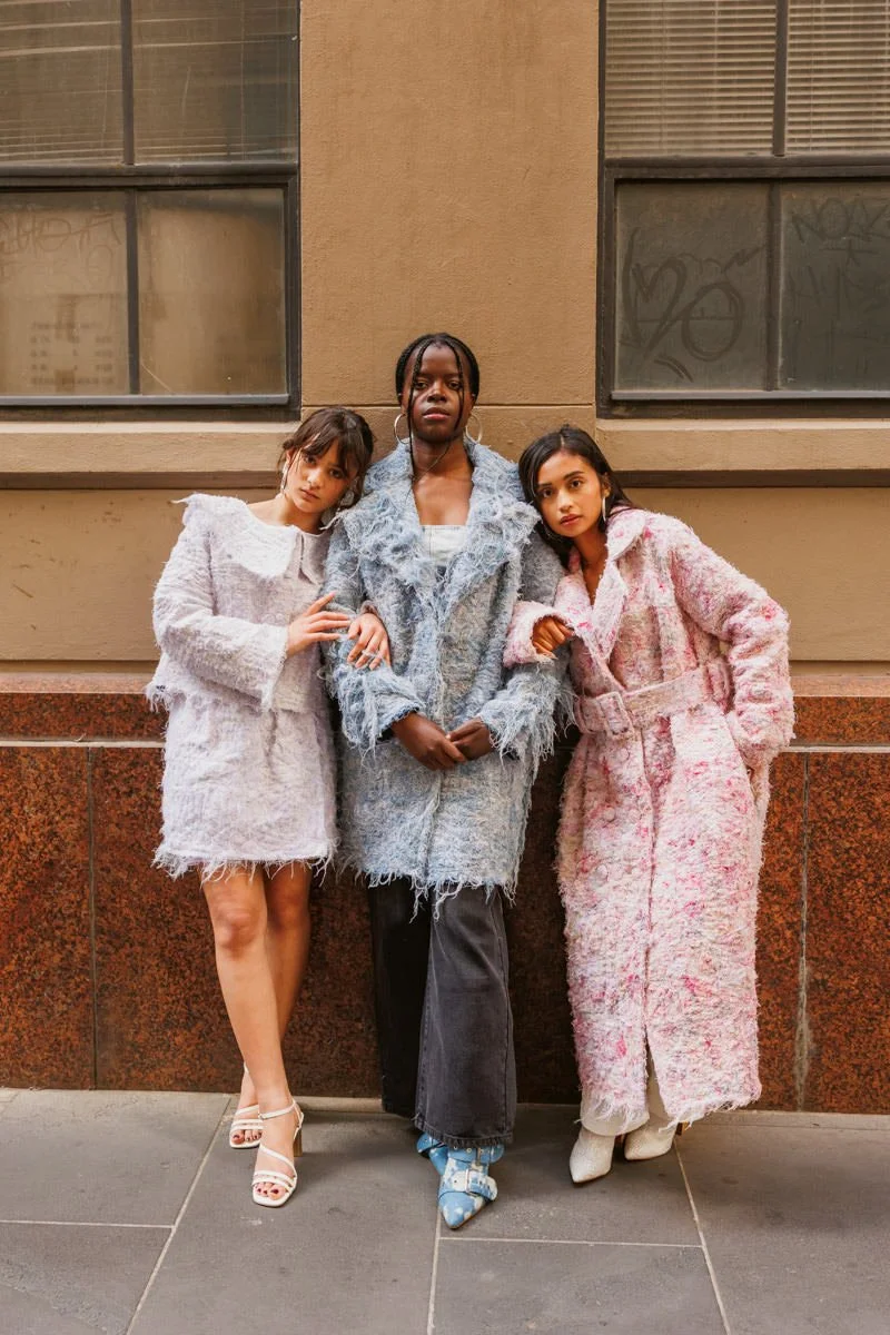 Three women standing side by side against a city building wall, wearing stylish textured coats, with the middle woman in black pants and the other two in dresses with high heels.