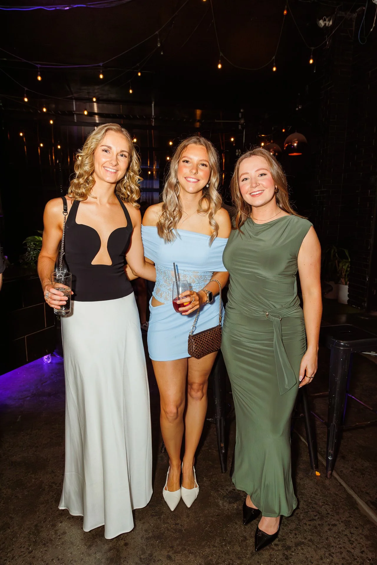 Three women in party dresses standing close together in a dimly lit indoor venue, smiling at the camera, holding drinks.