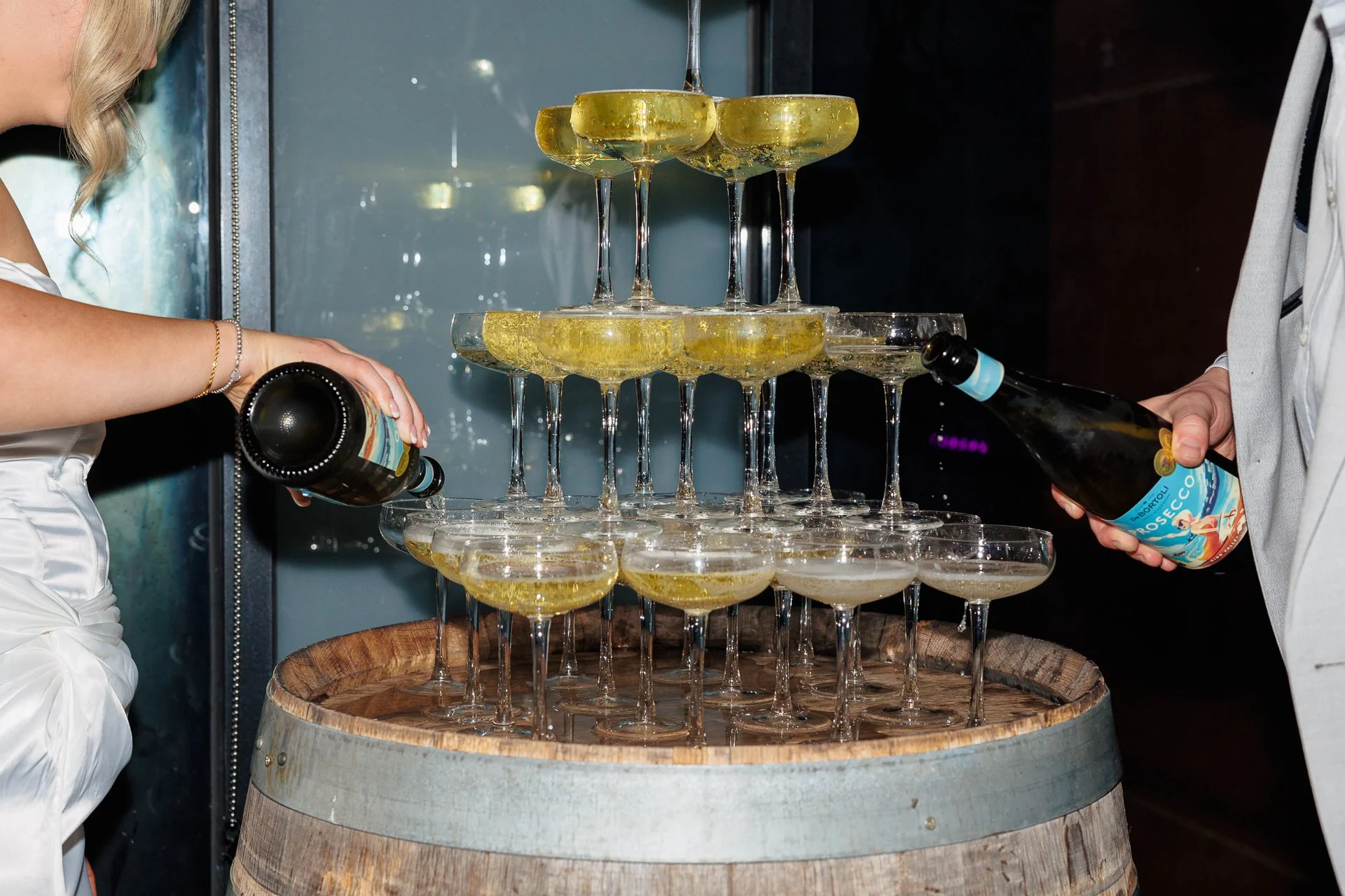 Two people pouring champagne into a pyramid of coupe glasses on top of a wooden barrel.