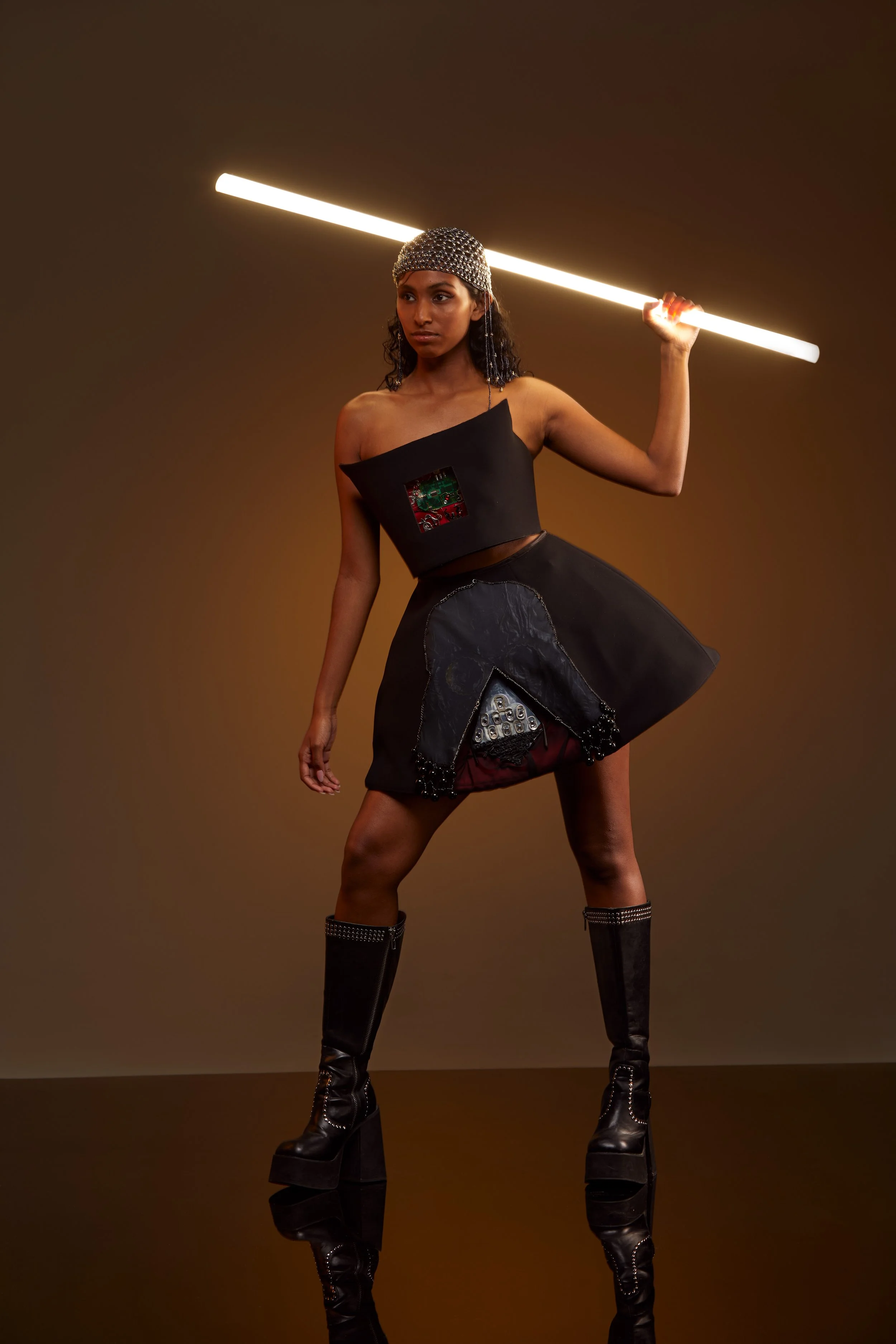 A woman wearing a futuristic black outfit with metallic details, a wide skirt, and knee-high platform boots, holding a glowing fluorescent tube light on her shoulder, standing against a plain brown background.