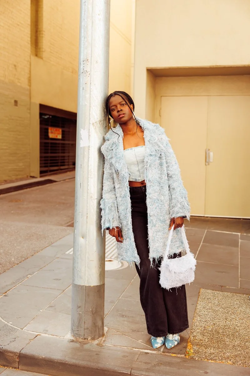 Young woman with braided hair standing against a pole on city sidewalk, wearing a fuzzy coat, crop top, black pants, and patterned shoes, holding a fuzzy white bag.