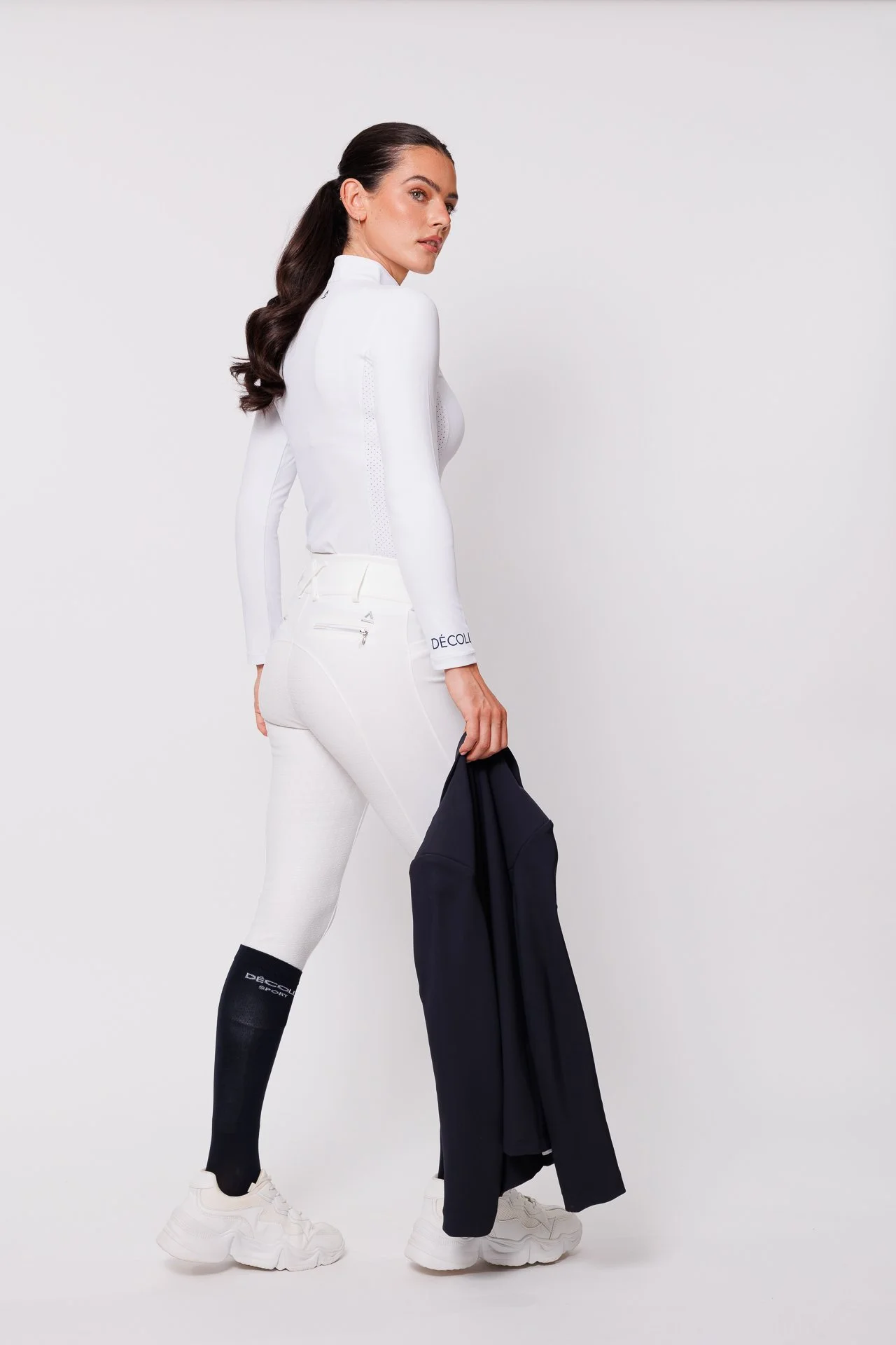 A woman with dark brown hair in loose curls, wearing a fitted white athletic jacket and white leggings, holding a dark jacket in her right hand, standing against a plain white background.