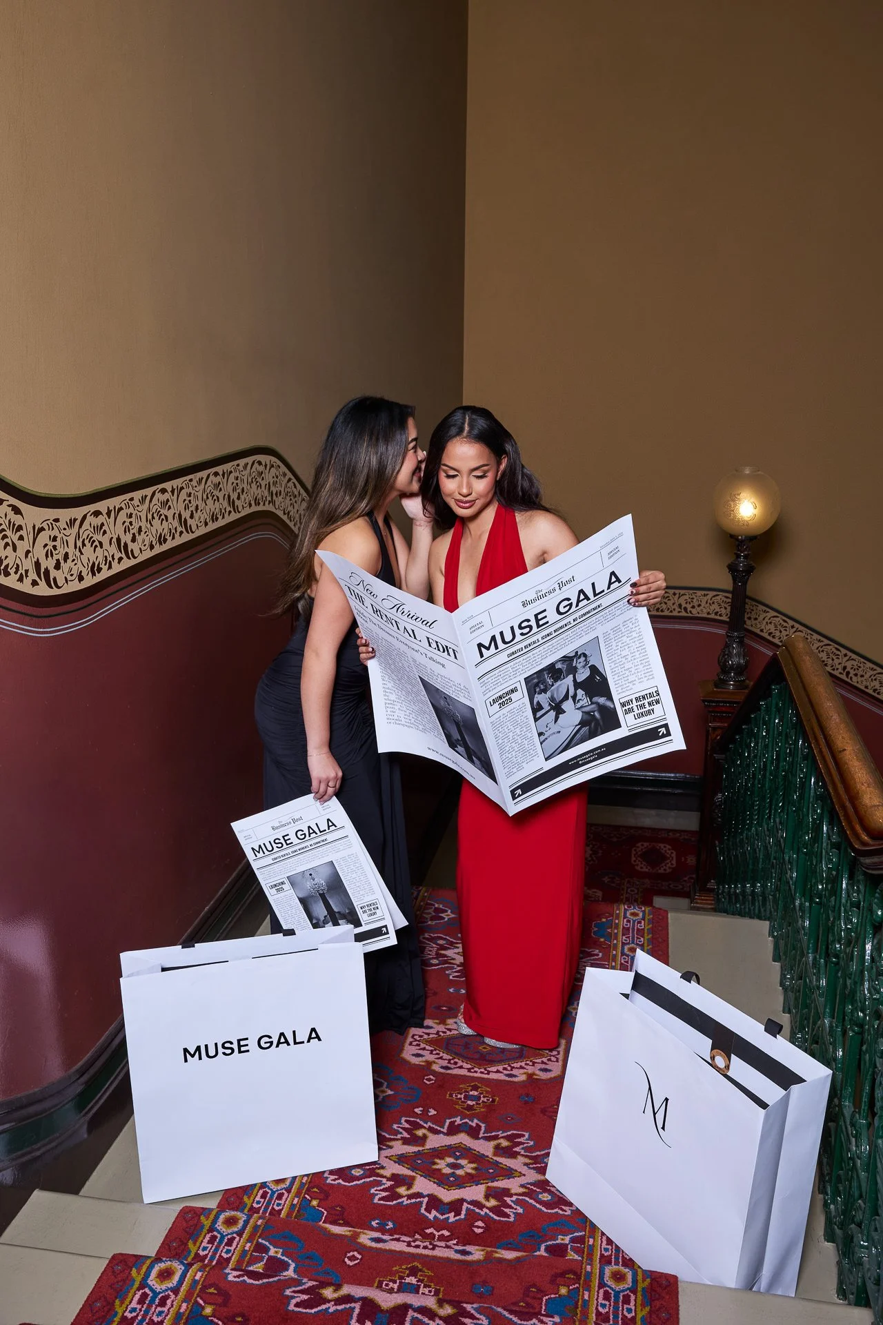 Two women in evening gowns reading a newspaper titled 'Muse Gala' on a stairway landing, with shopping bags labeled 'Muse Gala' nearby.
