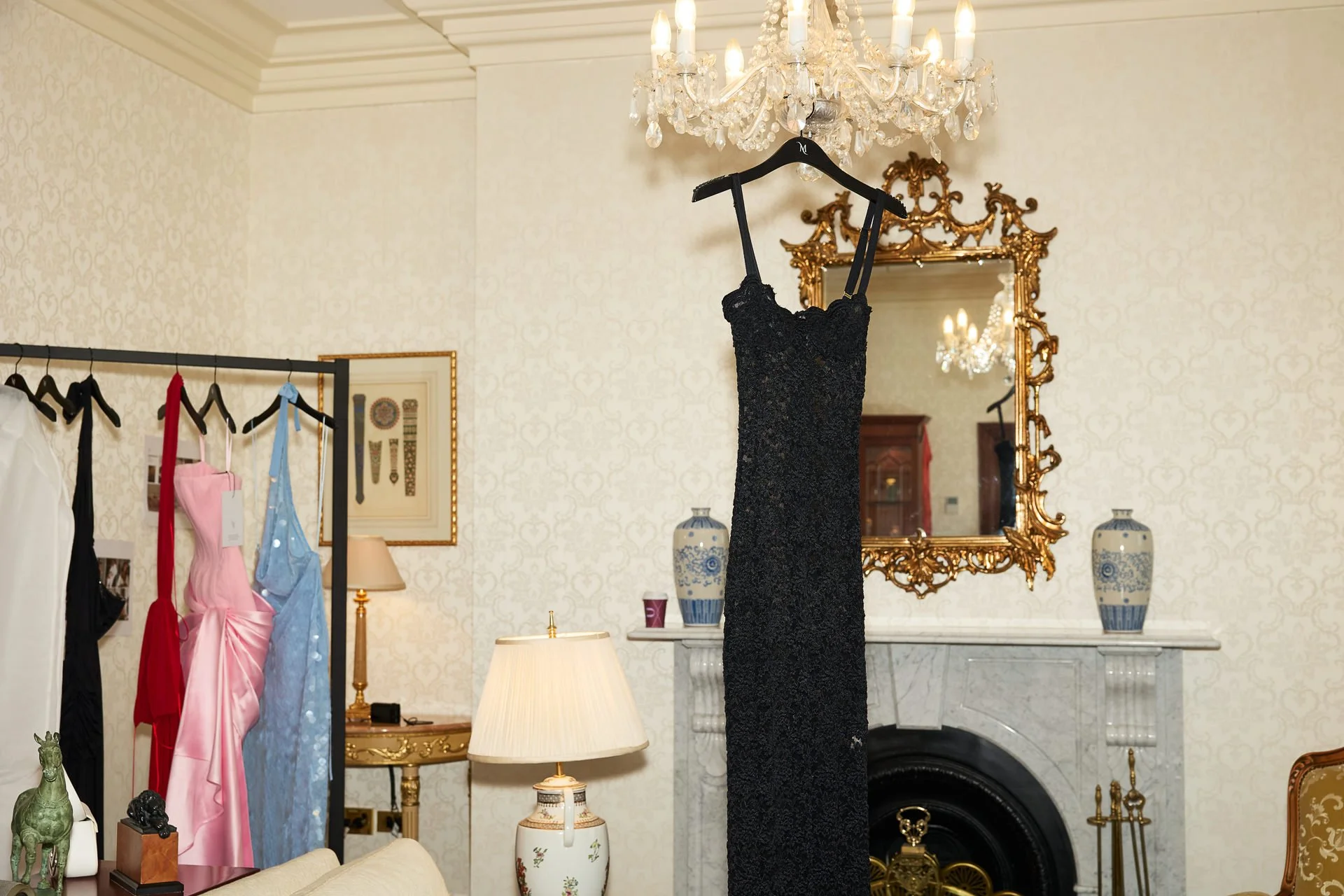 A black sequin dress hanging on a decorative black hanger in a room with ornate gold-framed mirror, marble fireplace, and pastel-colored dresses on a clothing rack.