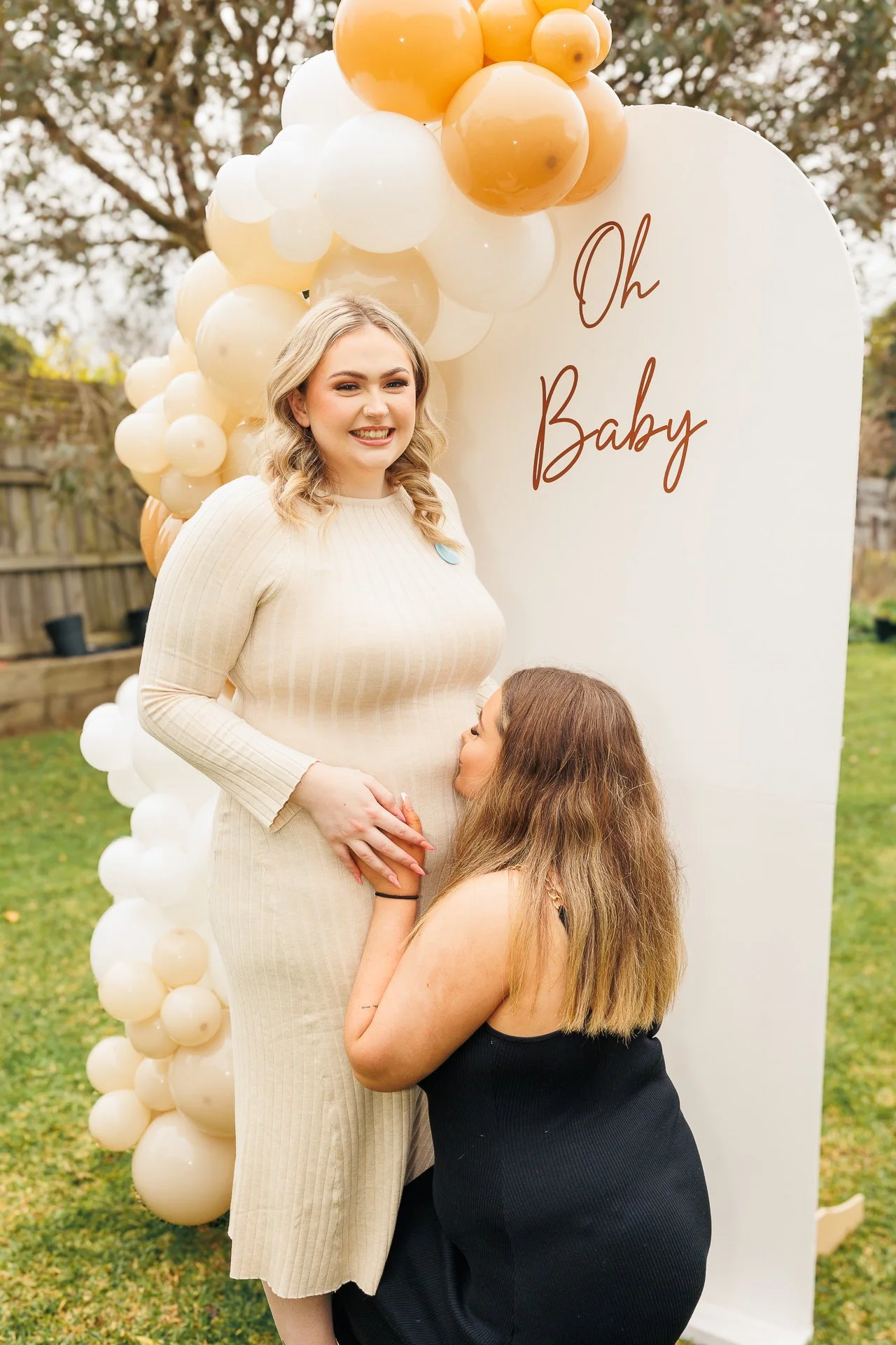 A pregnant woman in a beige dress smiling at the camera, being embraced affectionately by a woman in a black dress who is kneeling. They are outdoors near a white sign with 'Oh Baby' written on it and surrounded by beige and white balloons.