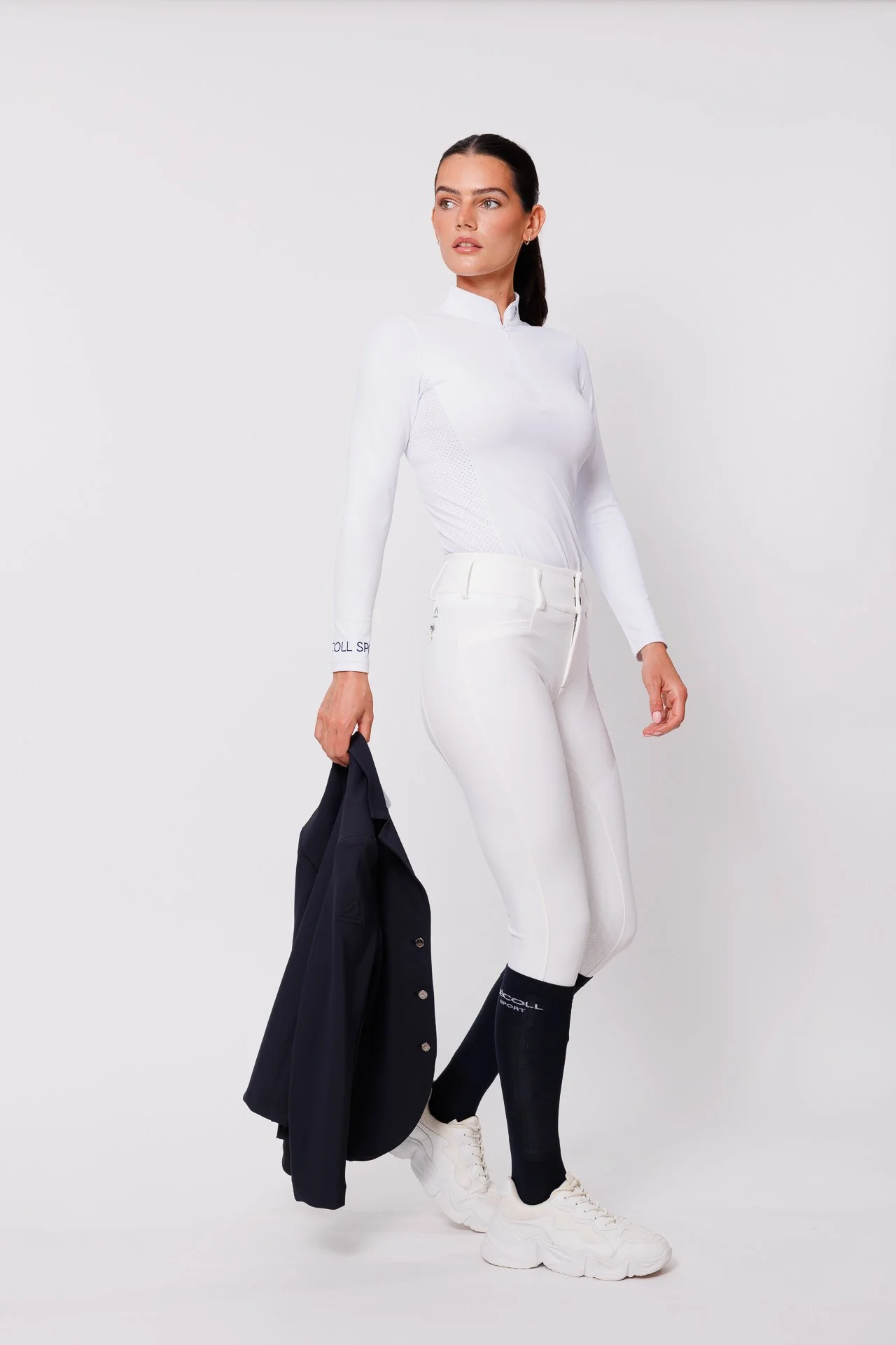 Woman in white athletic outfit holding navy blazer, standing on white background.