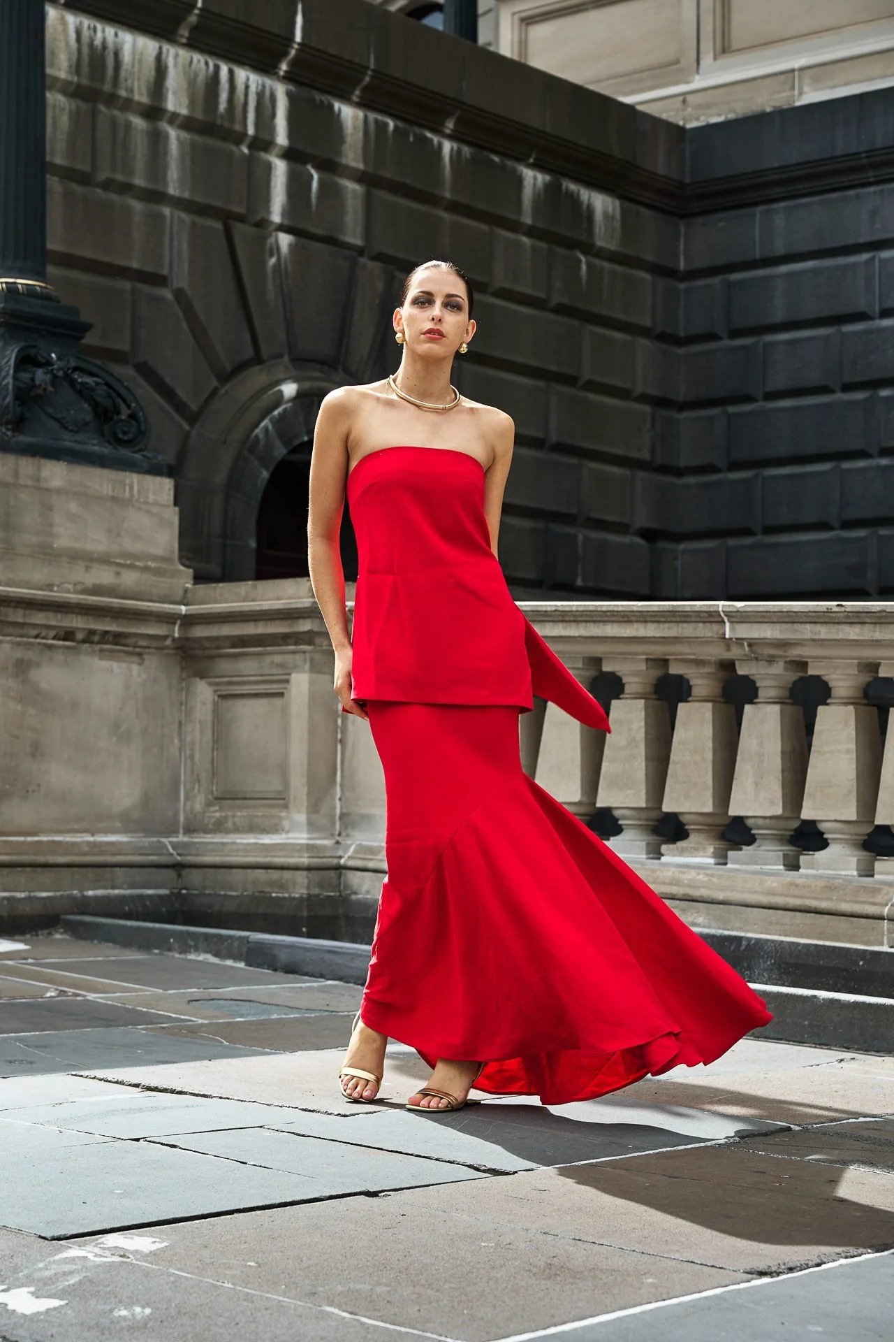 A woman in a strapless red dress stands on a city sidewalk with historic buildings in the background.