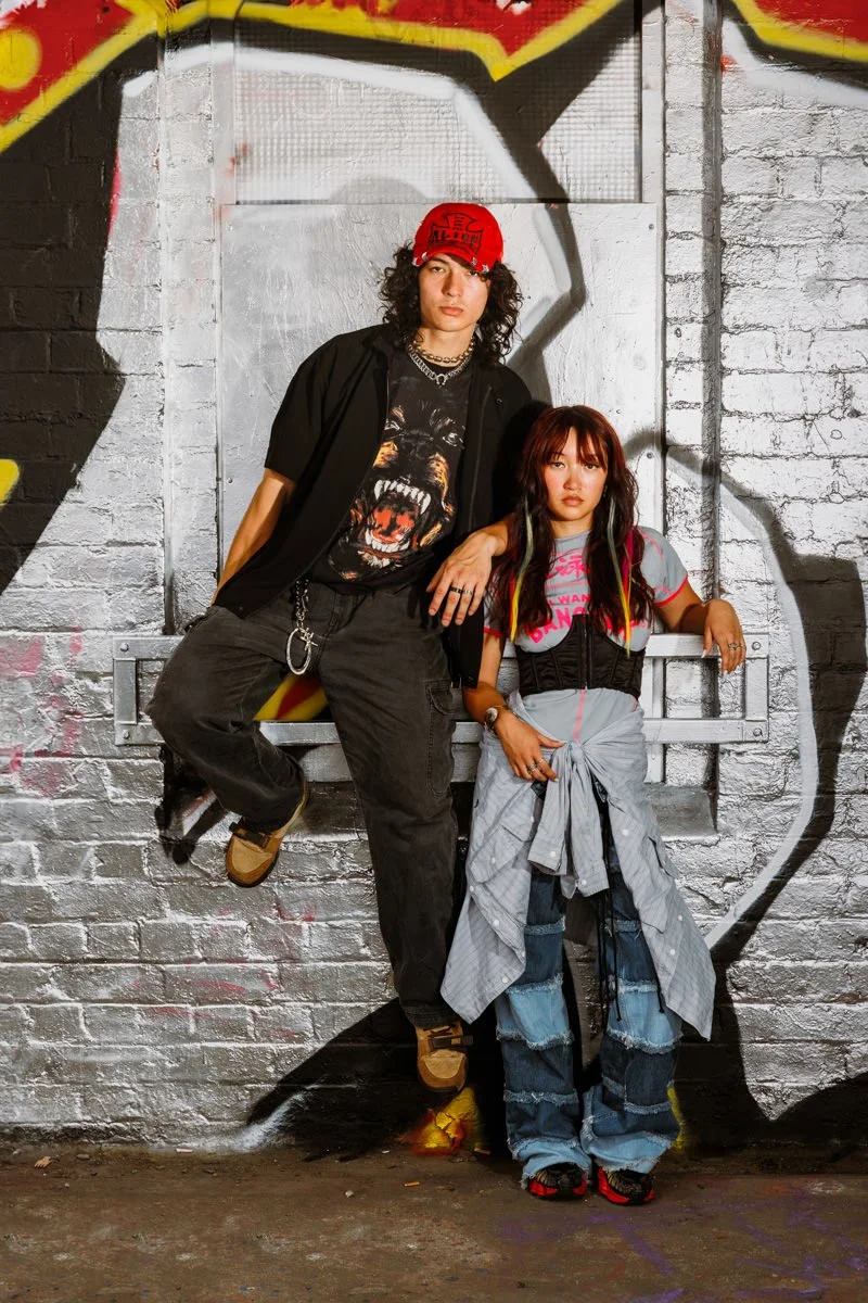 Two young people, one male and one female, pose in front of a graffiti-covered brick wall. The male, with long curly hair and a red cap, leans against the wall with his hand on the female's shoulder. The female has long, wavy hair with colorful strea