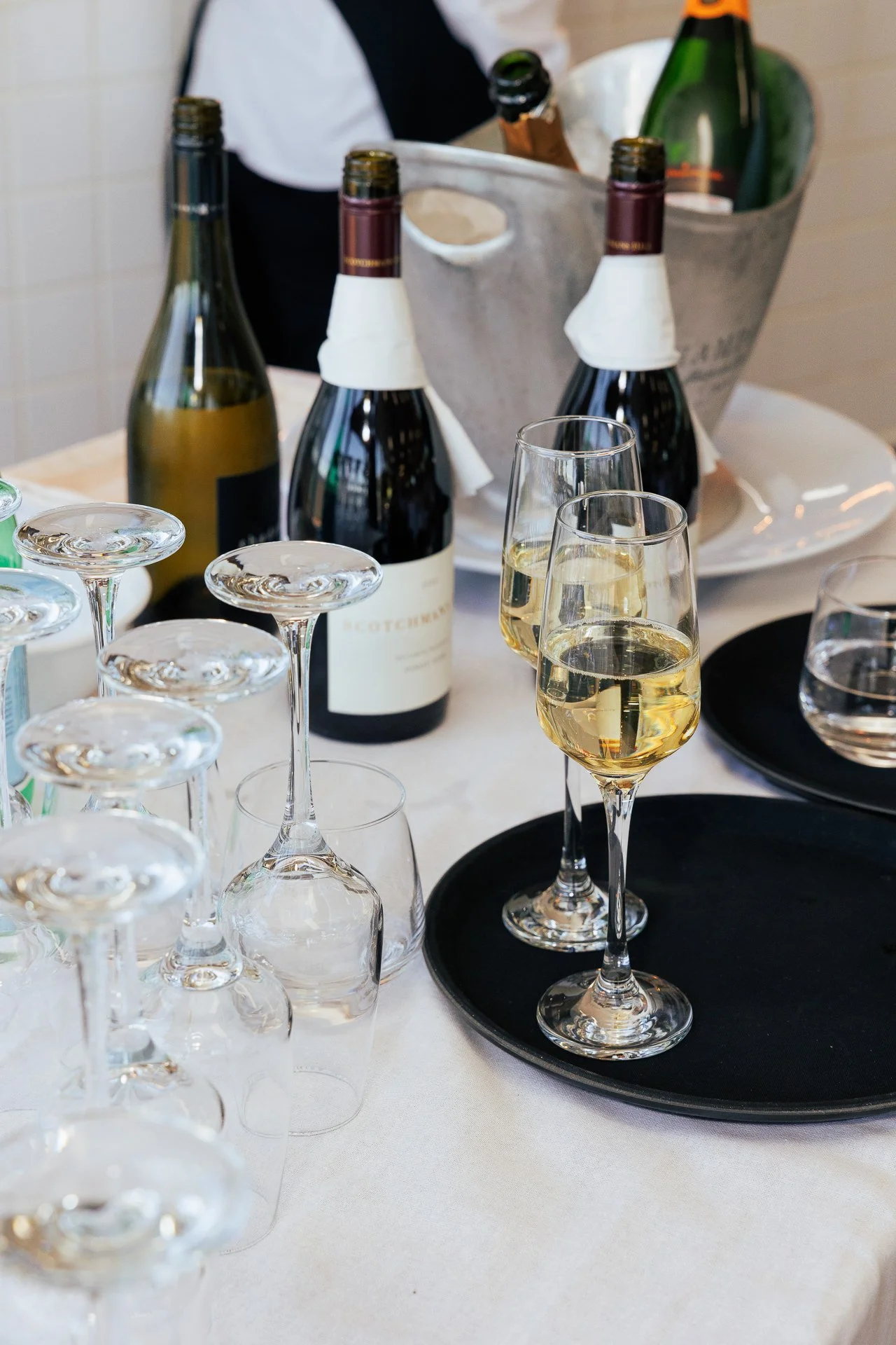 A table with multiple wine glasses, some filled with white wine, others upside down. Several bottles of wine are on the table, some with napkins around their necks. There is an ice bucket with more bottles in the background.