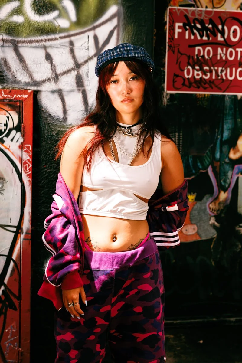 A young woman with dark hair, wearing a blue checkered cap, white crop top, purple camouflage pants, and a purple jacket, standing in front of graffiti-covered walls and signs.