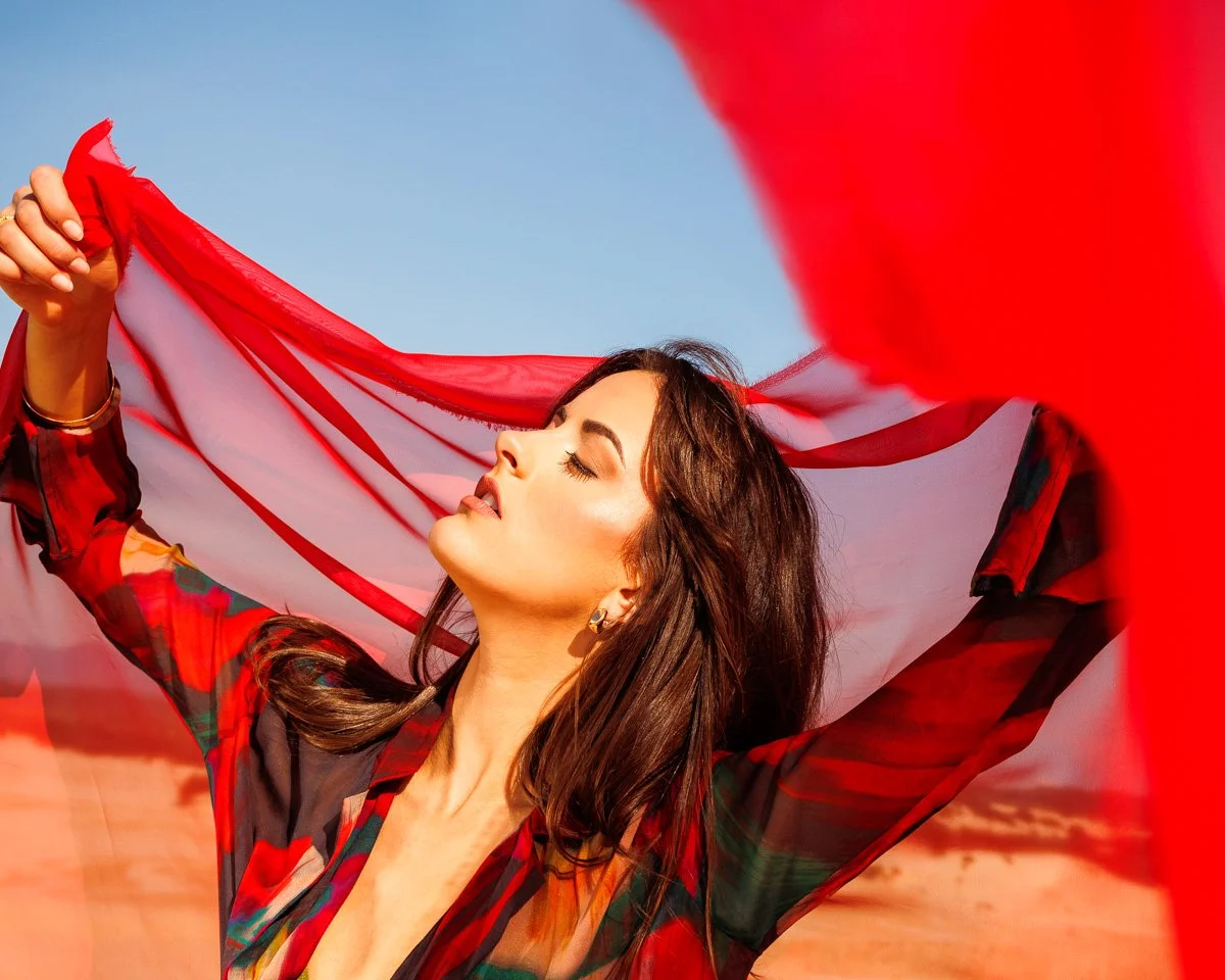A woman with dark brown hair and closed eyes holding a red and white flag or cloth up in the air outdoors against a blue sky.