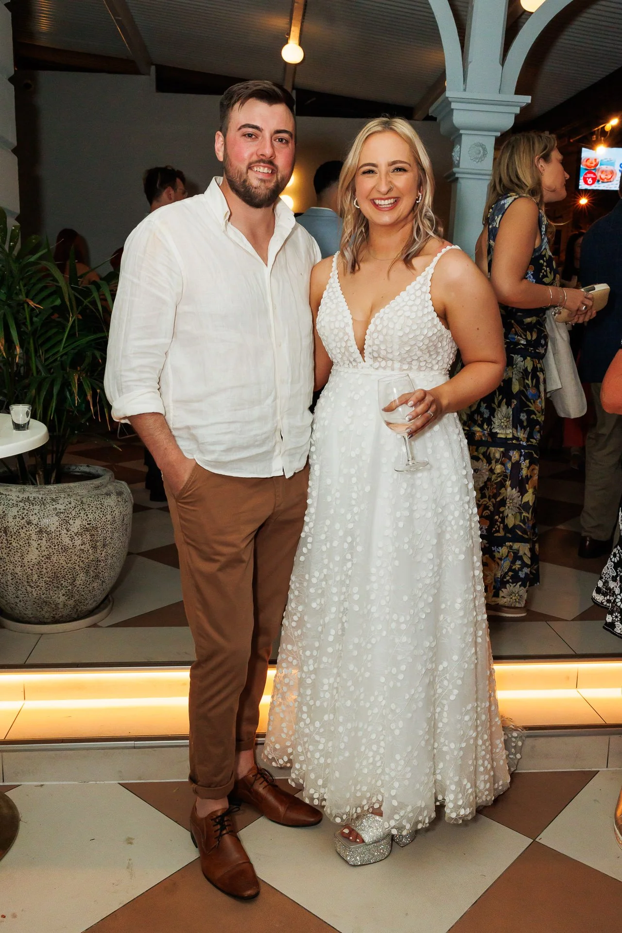 A man and woman posing at an indoor event, with the woman wearing a white dress and holding a glass of wine, and the man wearing a white shirt and brown pants.