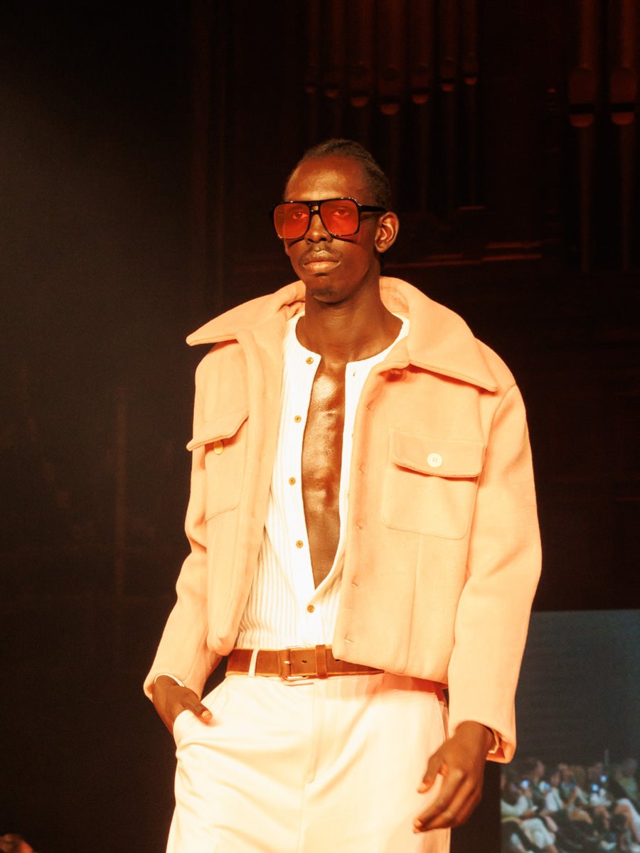 Fashion model walking on runway, wearing a peach-colored jacket over an open white shirt, beige pants, sunglasses, and a belt.