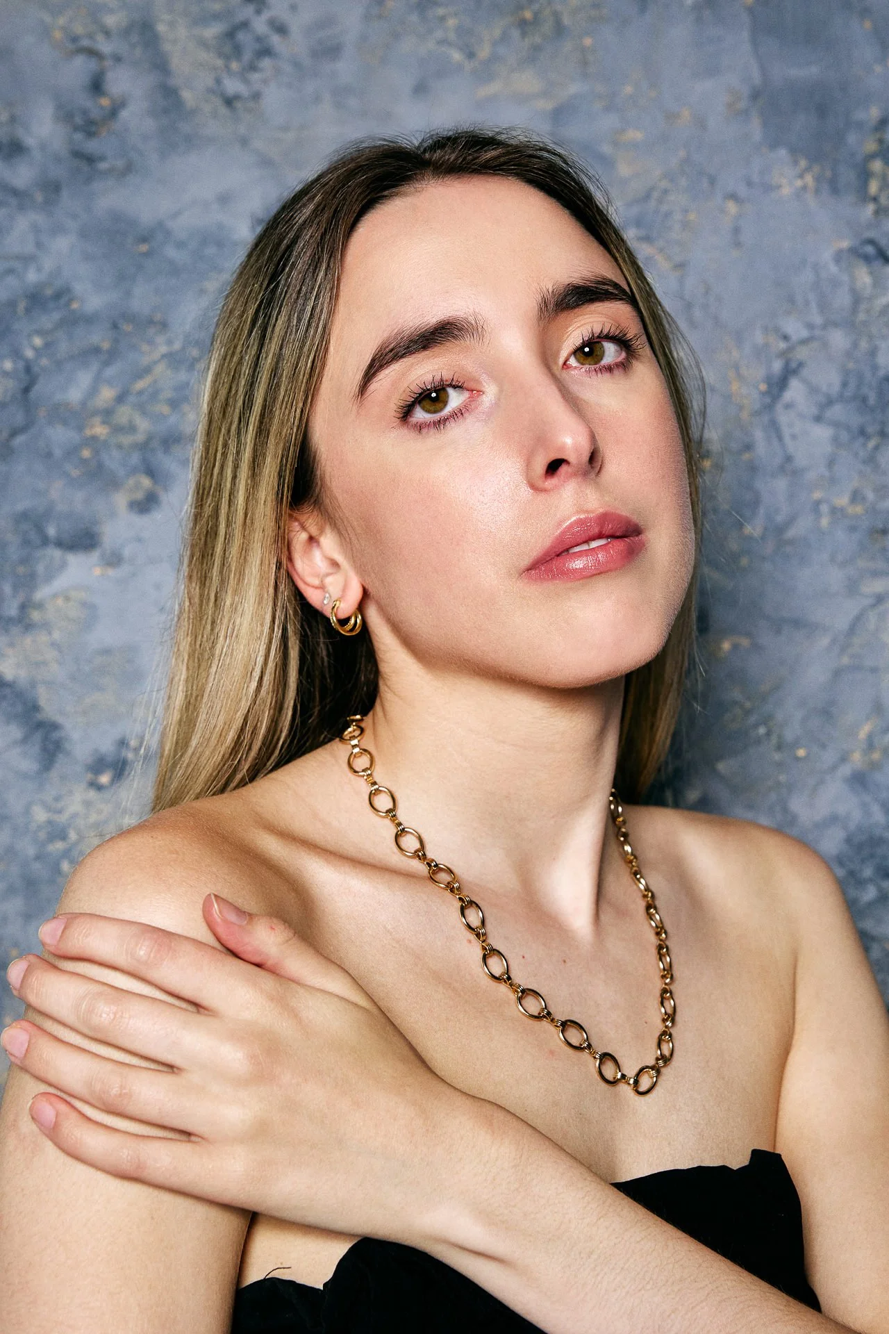 A young woman with long blonde hair, green eyes, and natural makeup, wearing gold jewelry, with a textured gray-blue background.