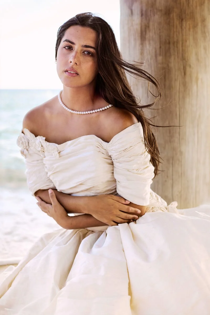 A woman in a white off-shoulder dress sitting outdoors near a large wooden structure by the beach, with ocean in the background.